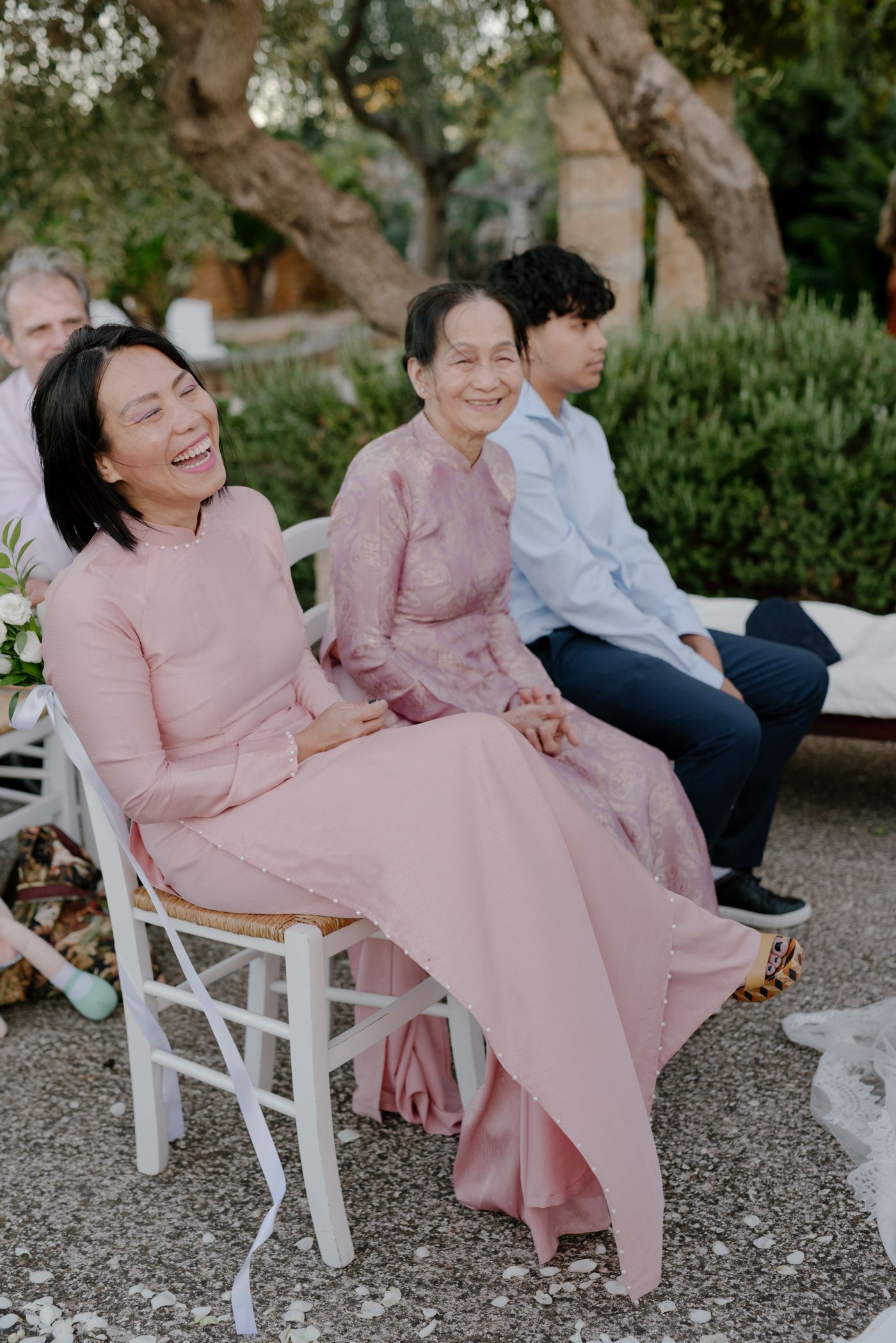 Wedding guests in matching pink dresses share joyful laughter while seated on white chairs at an outdoor event.