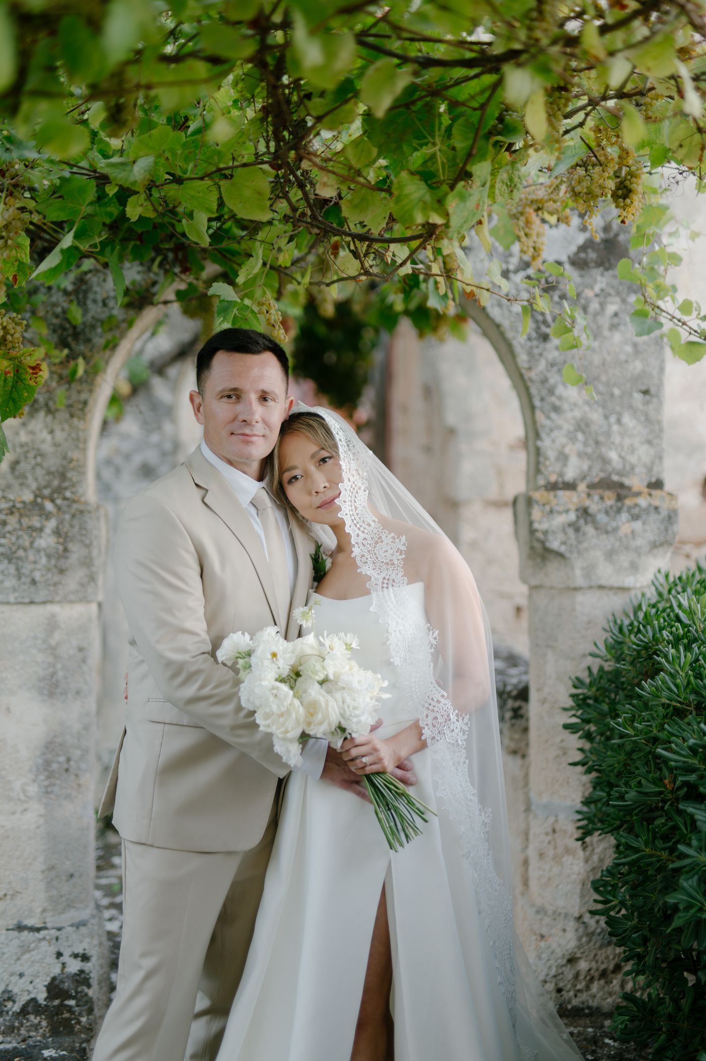 A wedding couple poses together in a romantic garden setting with stone walls and green foliage in the background.