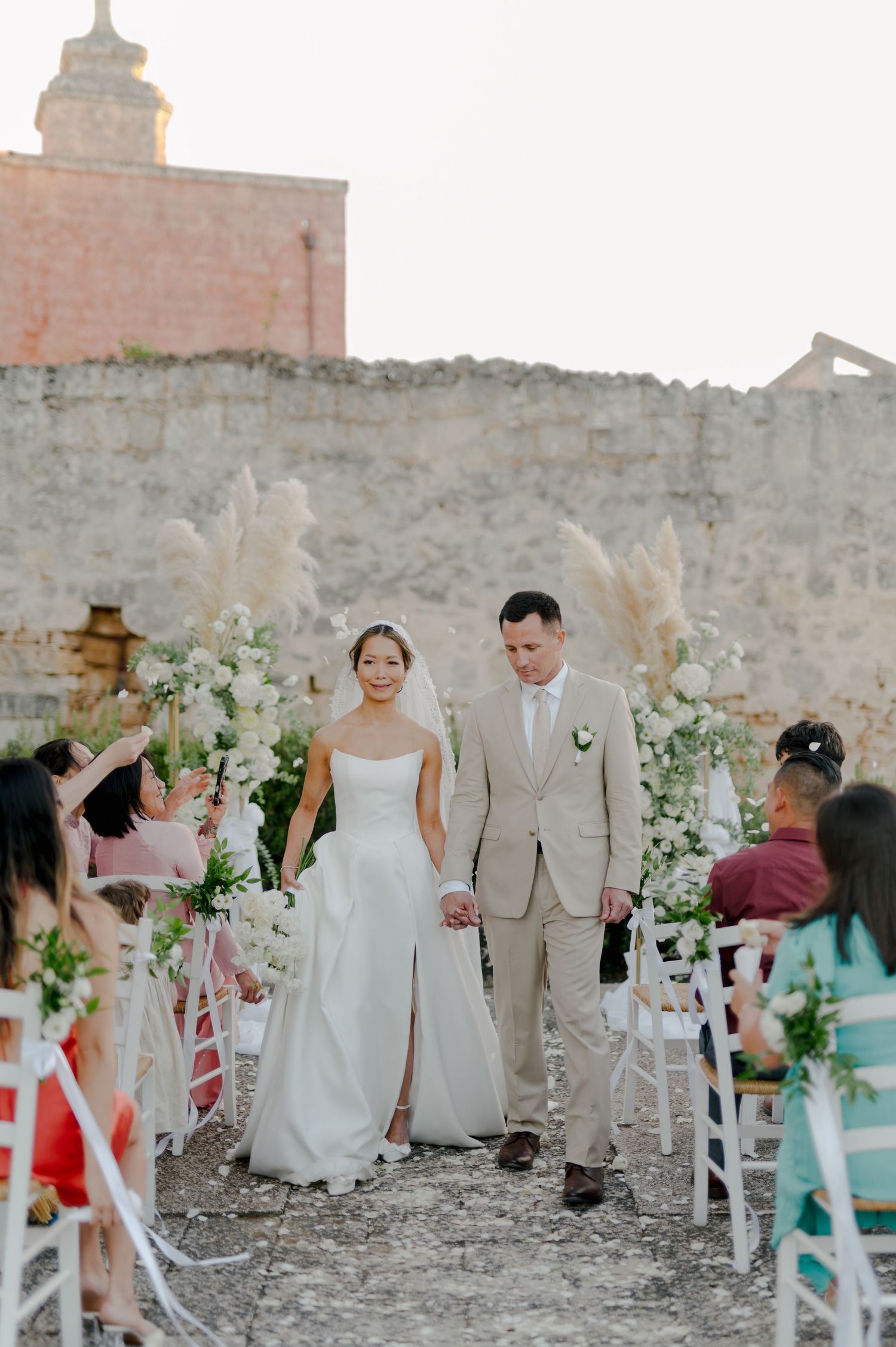 Wedding party walking down aisle decorated with white flowers at historic stone venue during outdoor ceremony.