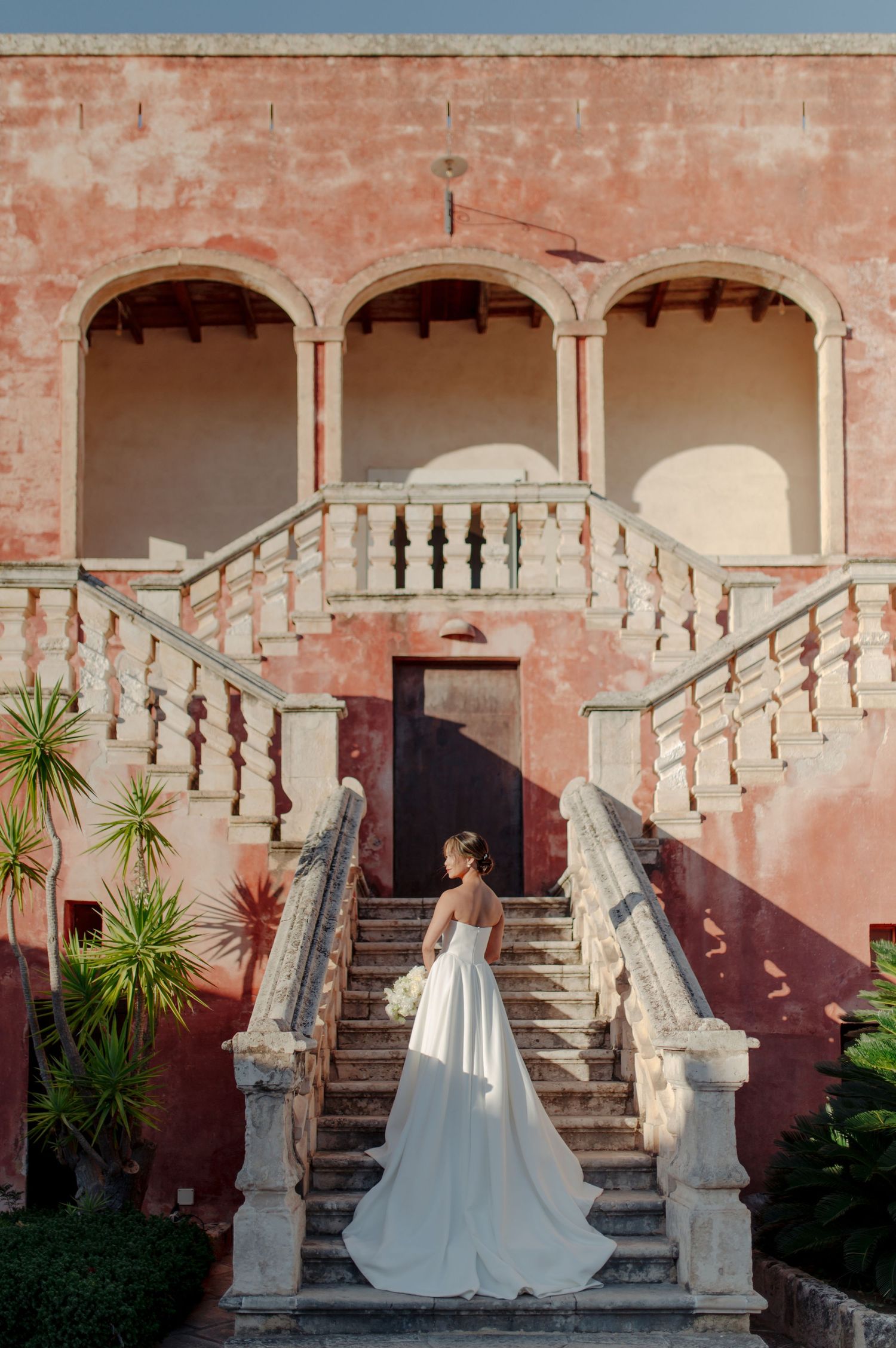 A bride in a flowing white gown poses on ornate stone steps against a pink stucco wall with classical architectural details.