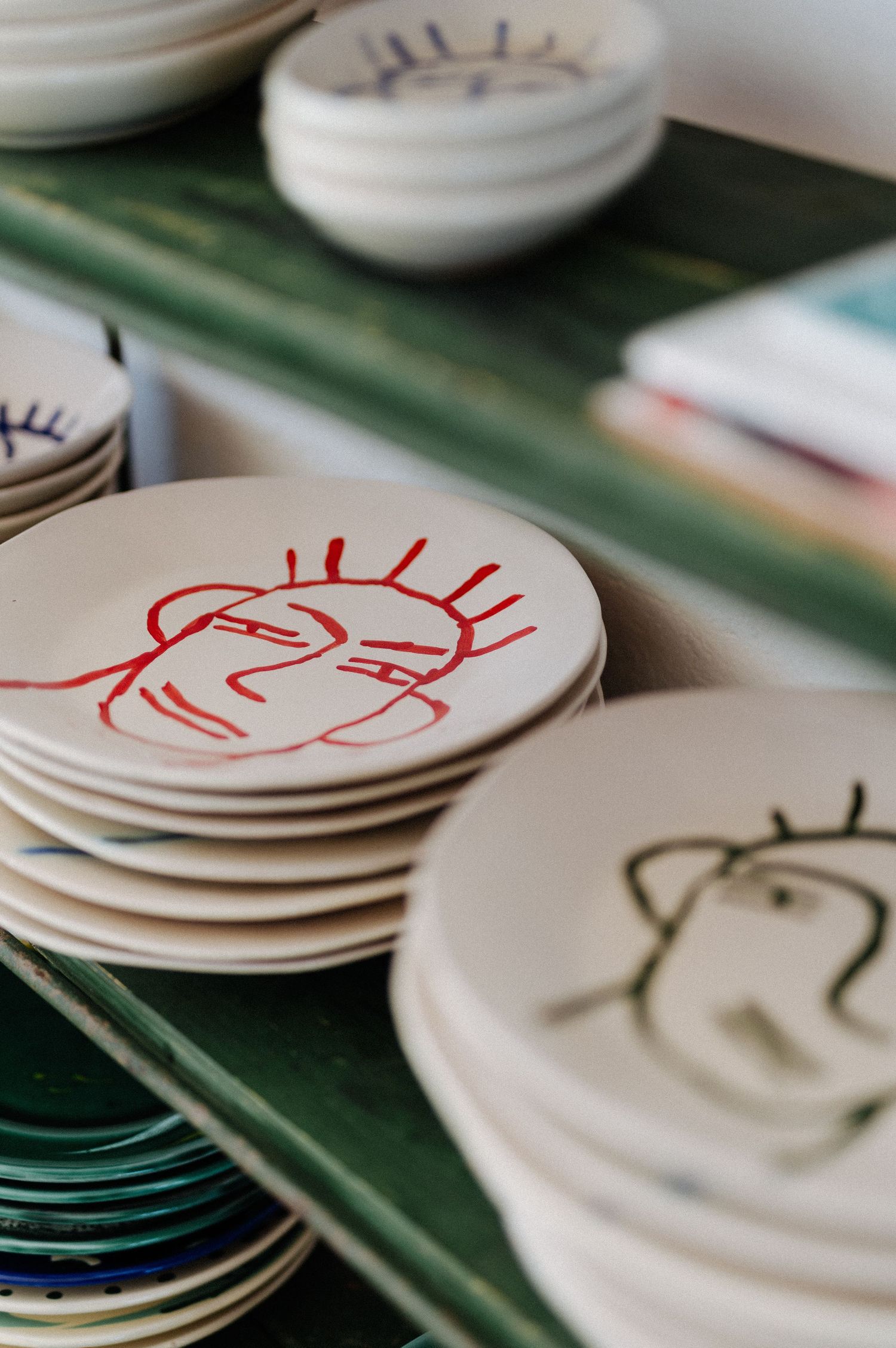 Stacks of white ceramic plates with minimalist line drawings in red, black and blue on a green surface.