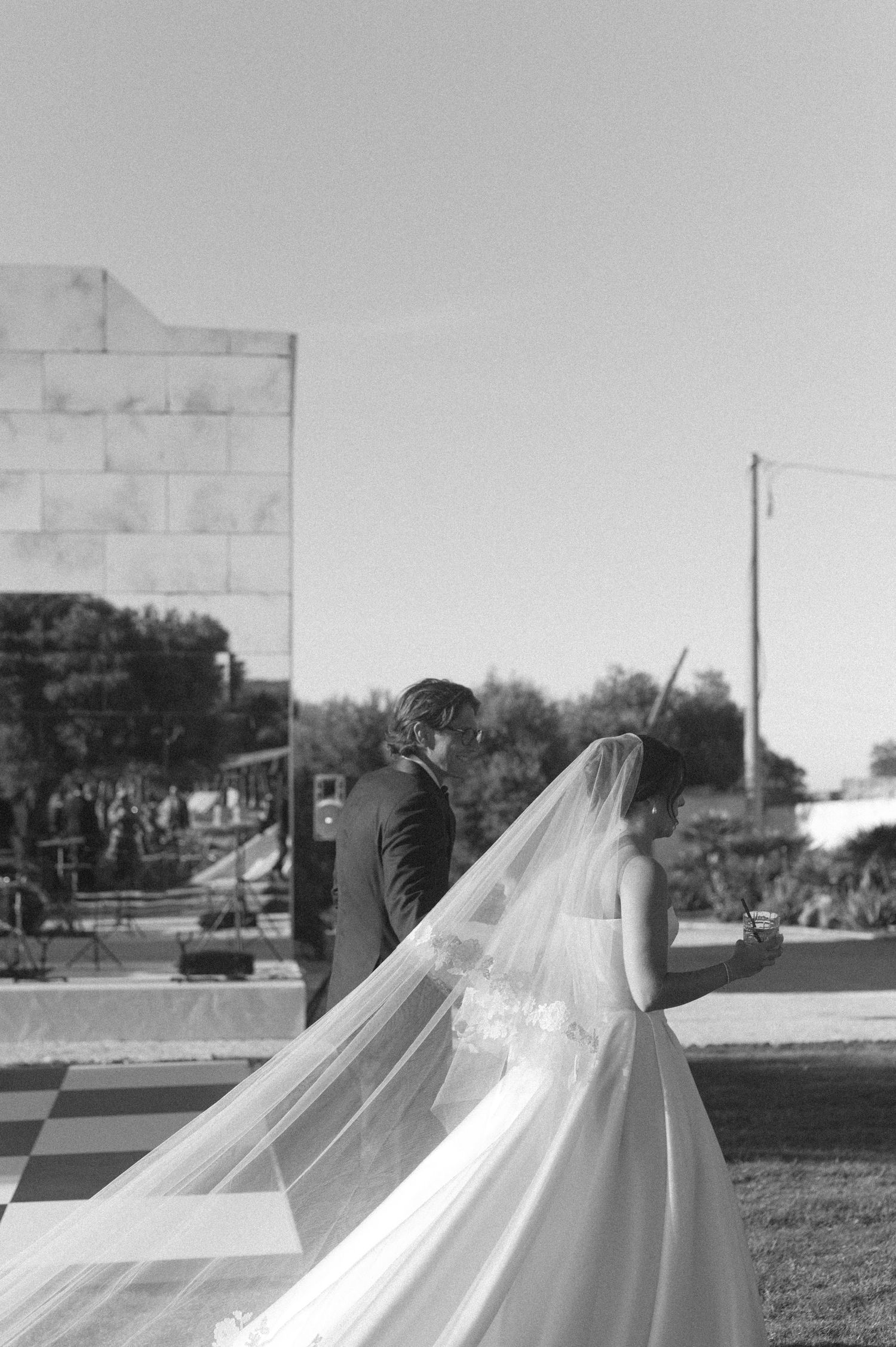 Black and white wedding photo of bride and groom walking outdoors, her long veil flowing gracefully in the breeze.