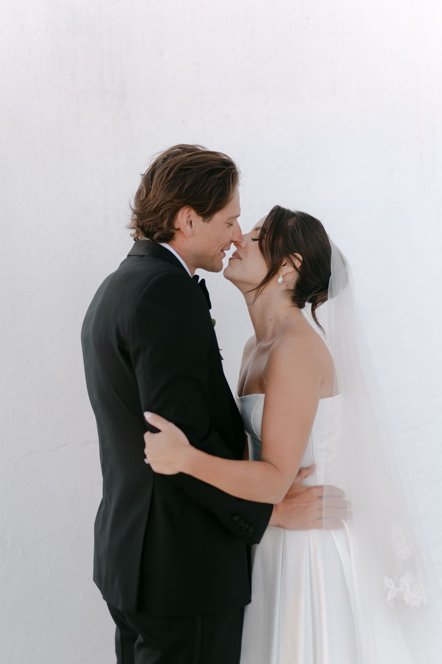 Bride and groom sharing a romantic almost-kiss, embracing closely against a bright white wall.