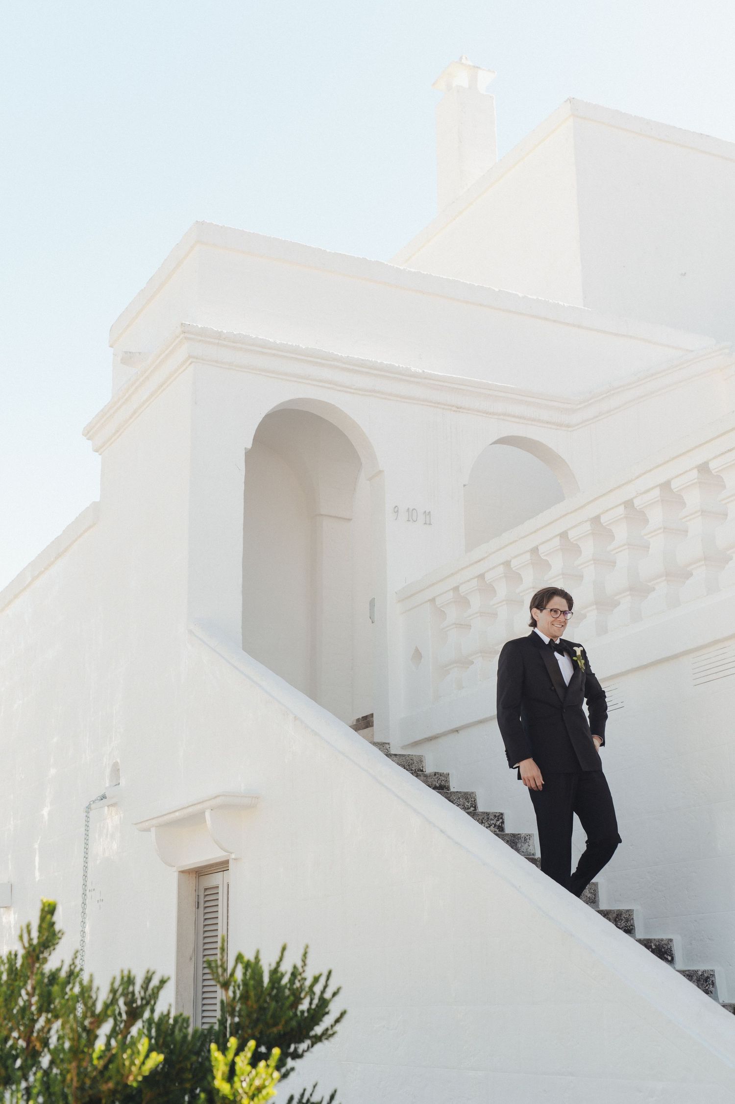 Groom in black tuxedo standing on white staircase of elegant Mediterranean-style venue under clear sky.