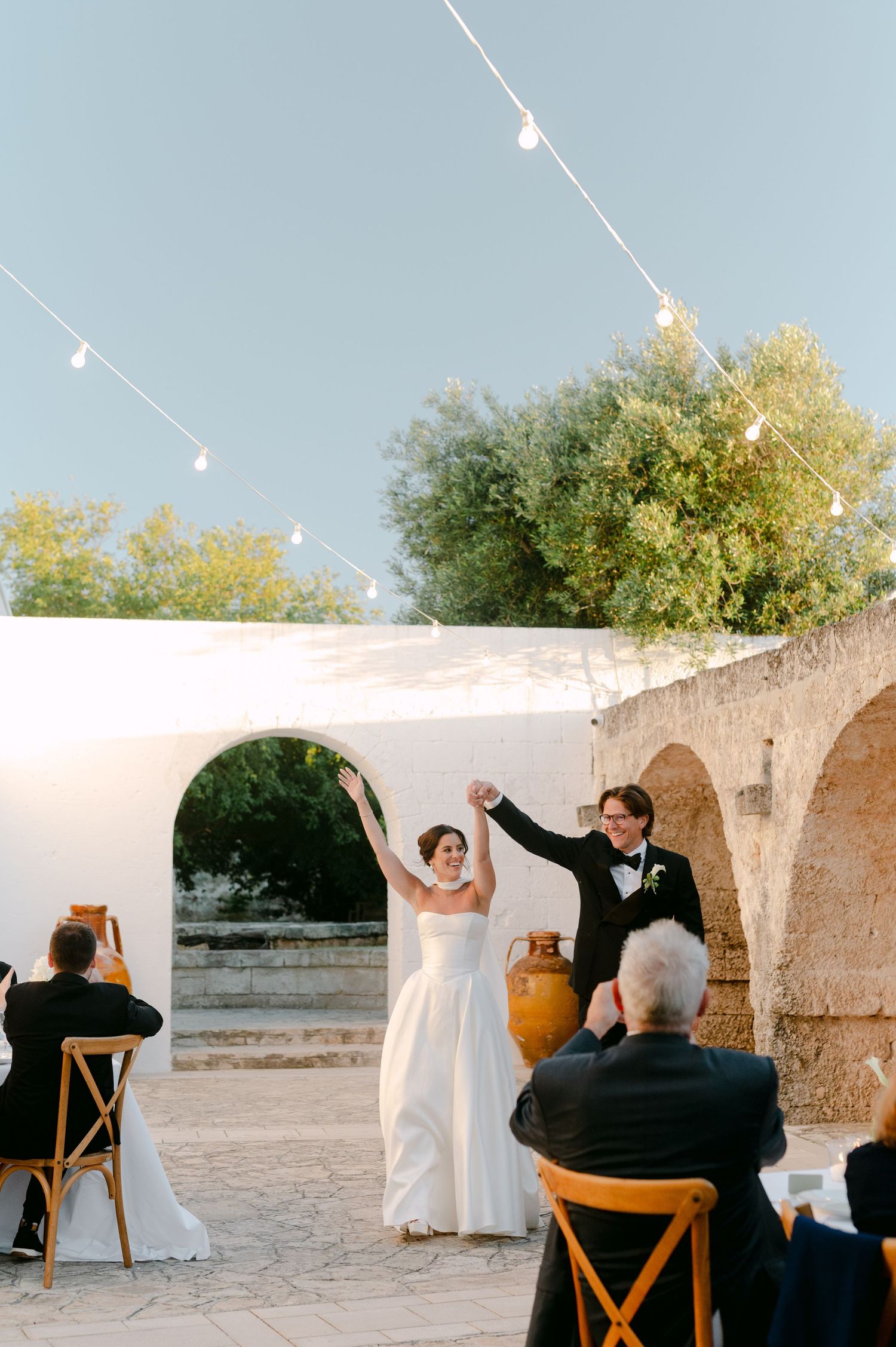Bride and groom raise hands in celebration at outdoor wedding ceremony with string lights and white arched wall.