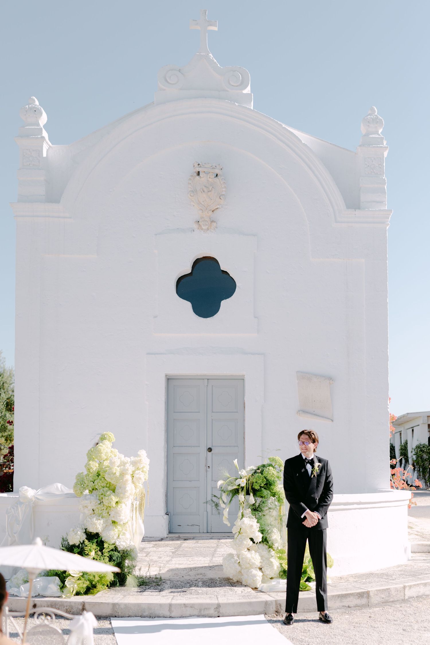 Groom in black suit stands before a white Mediterranean chapel adorned with white floral arrangements on a sunny wedding day.