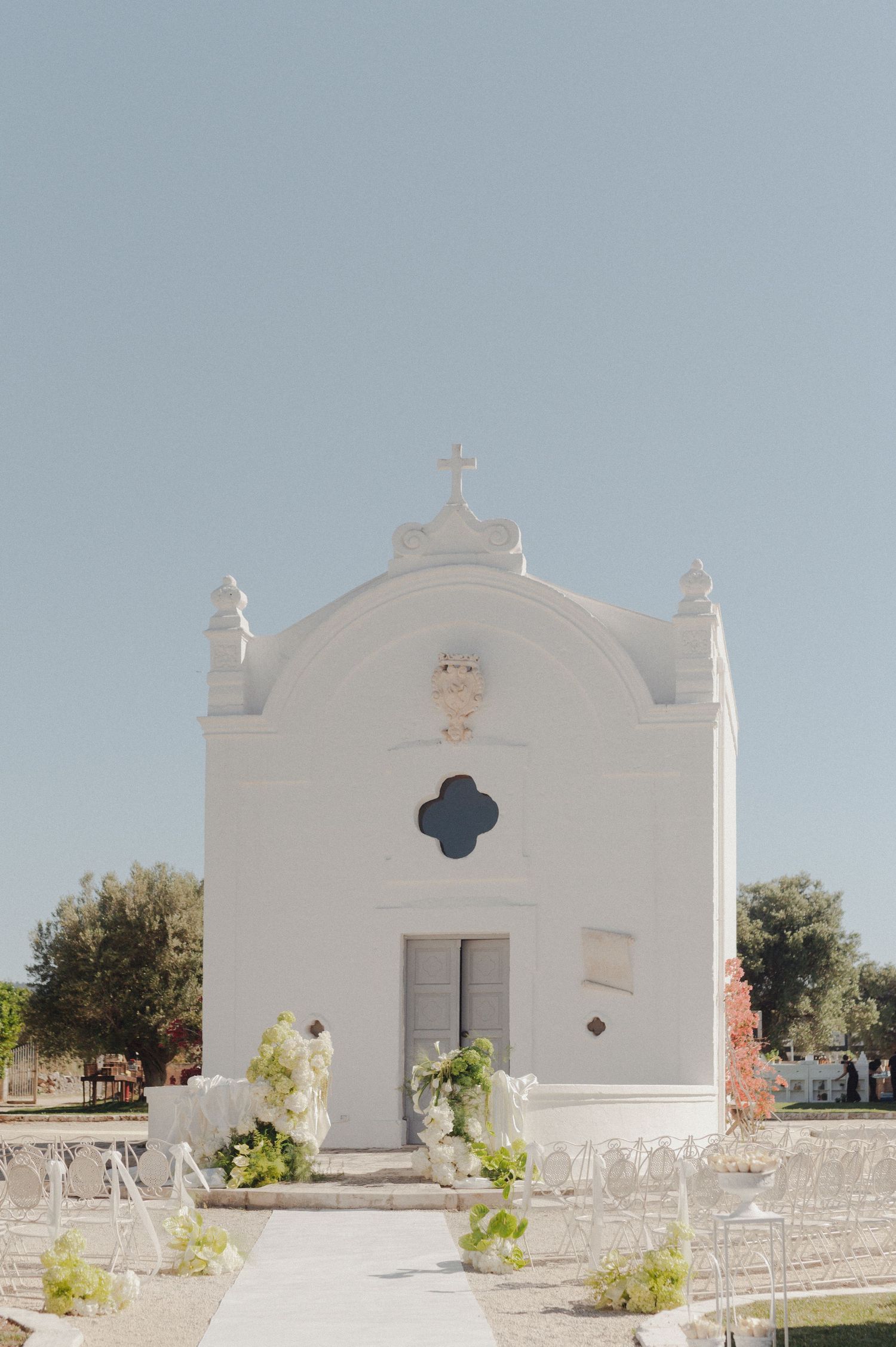 White chapel wedding ceremony setup with floral arrangements and white chairs on a sunny day.