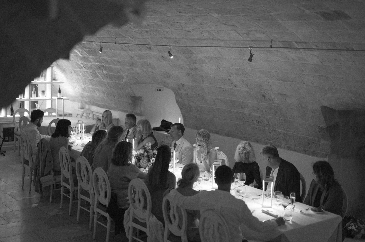 Black and white panorama of guests seated at a long dining table in a vaulted cellar.