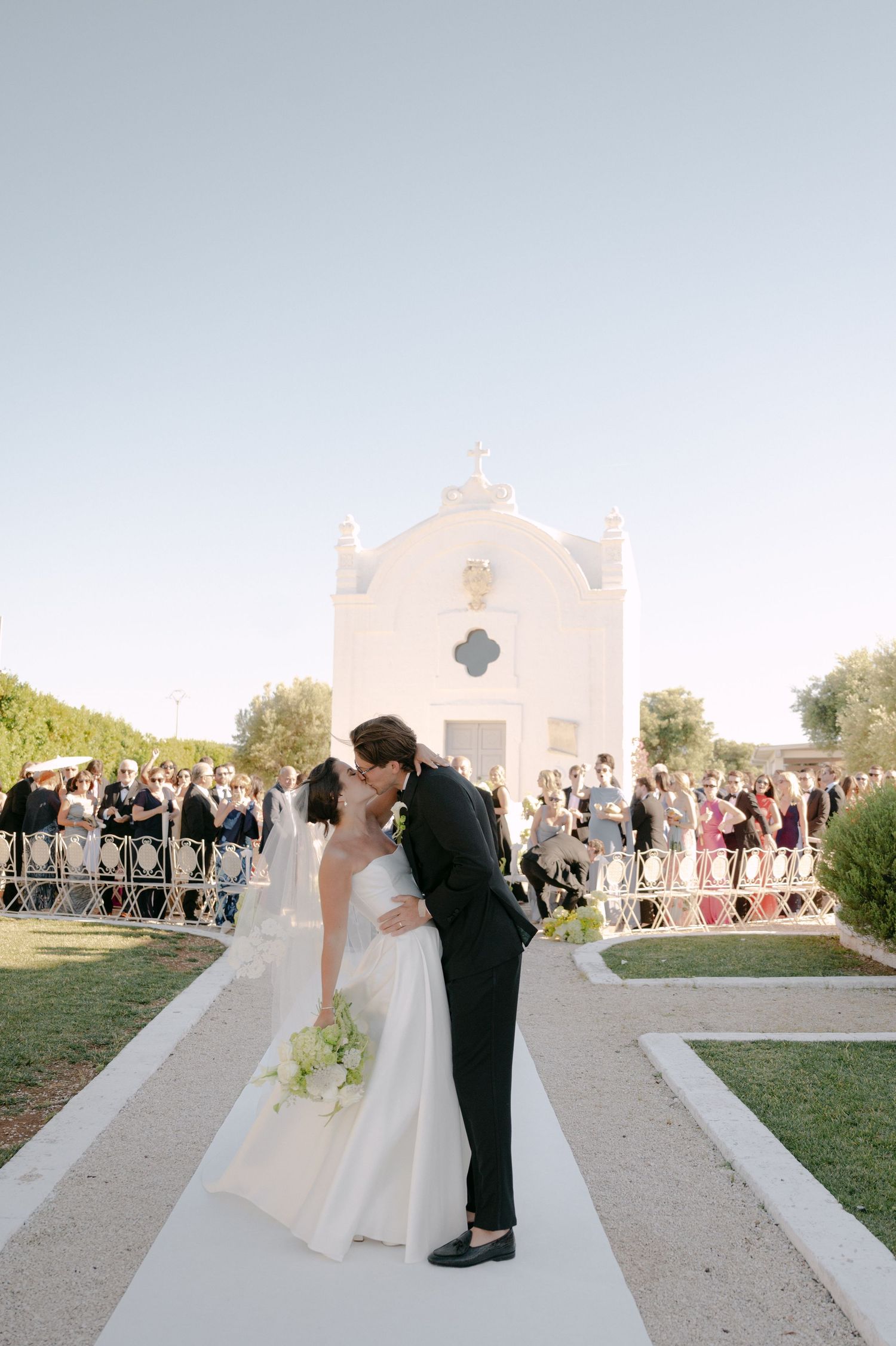 Bride and groom share first kiss on white pathway outside a white chapel with wedding guests gathered behind them.