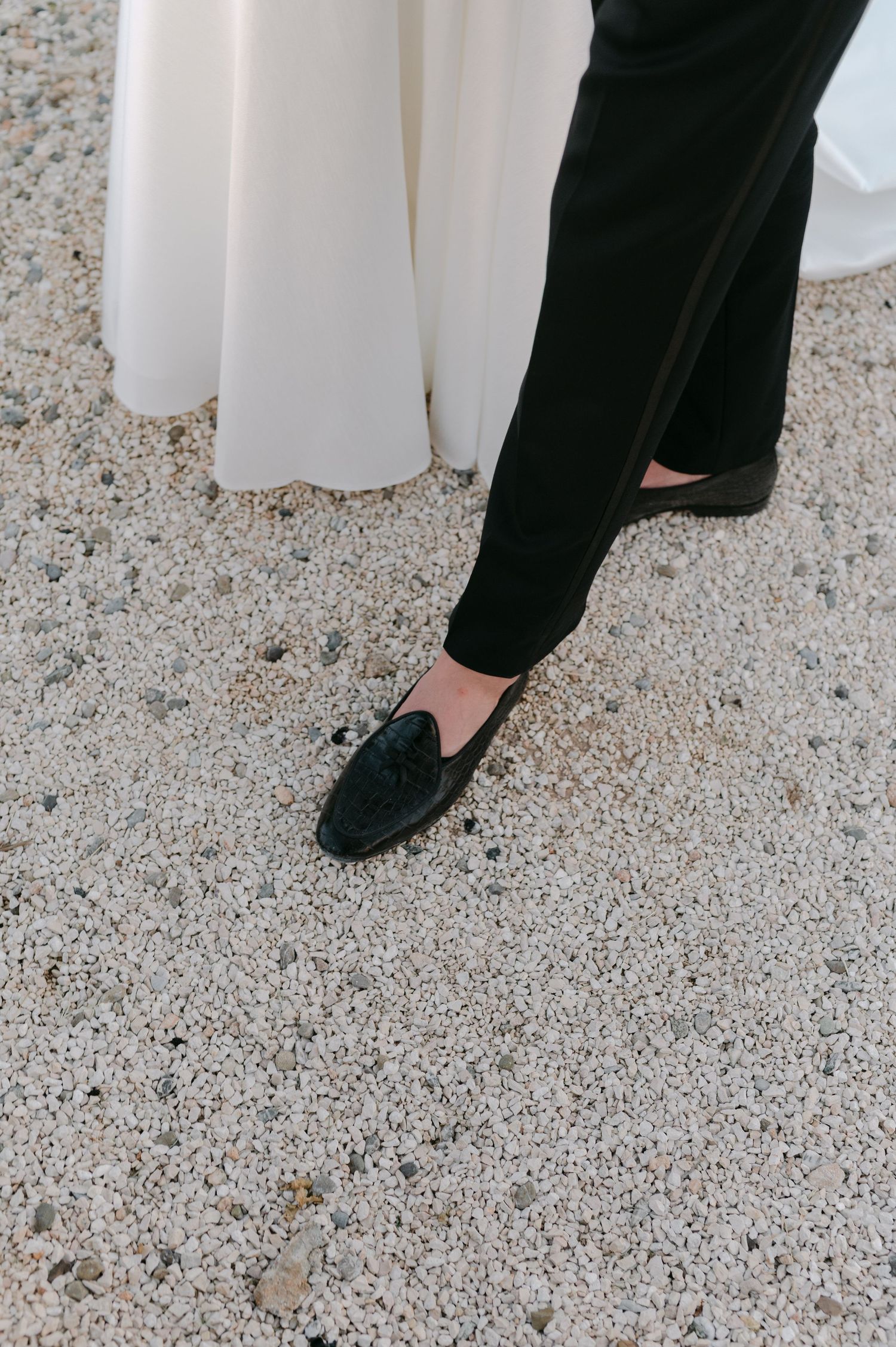 Close-up of groom's black tassel loafer and black tuxedo pants beside bride's white wedding dress on gravel.