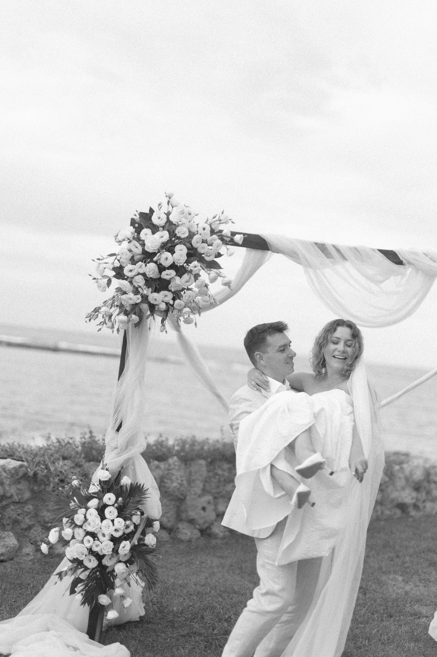 Romantic wedding ceremony setup with floral arch and flowing white fabric by the sea.
