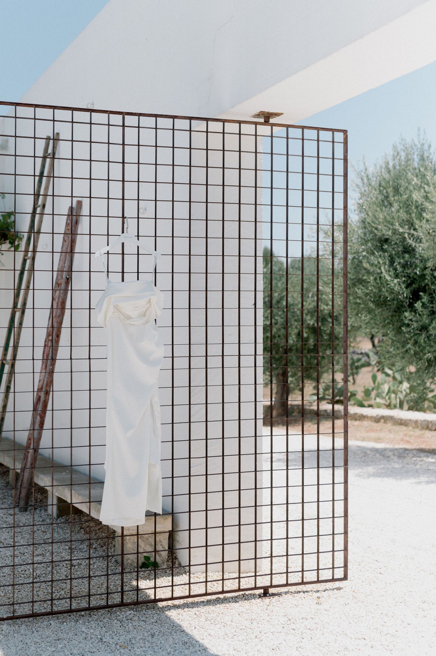 A white wedding dress hangs on a metal grid display panel outdoors with olive trees in the background.