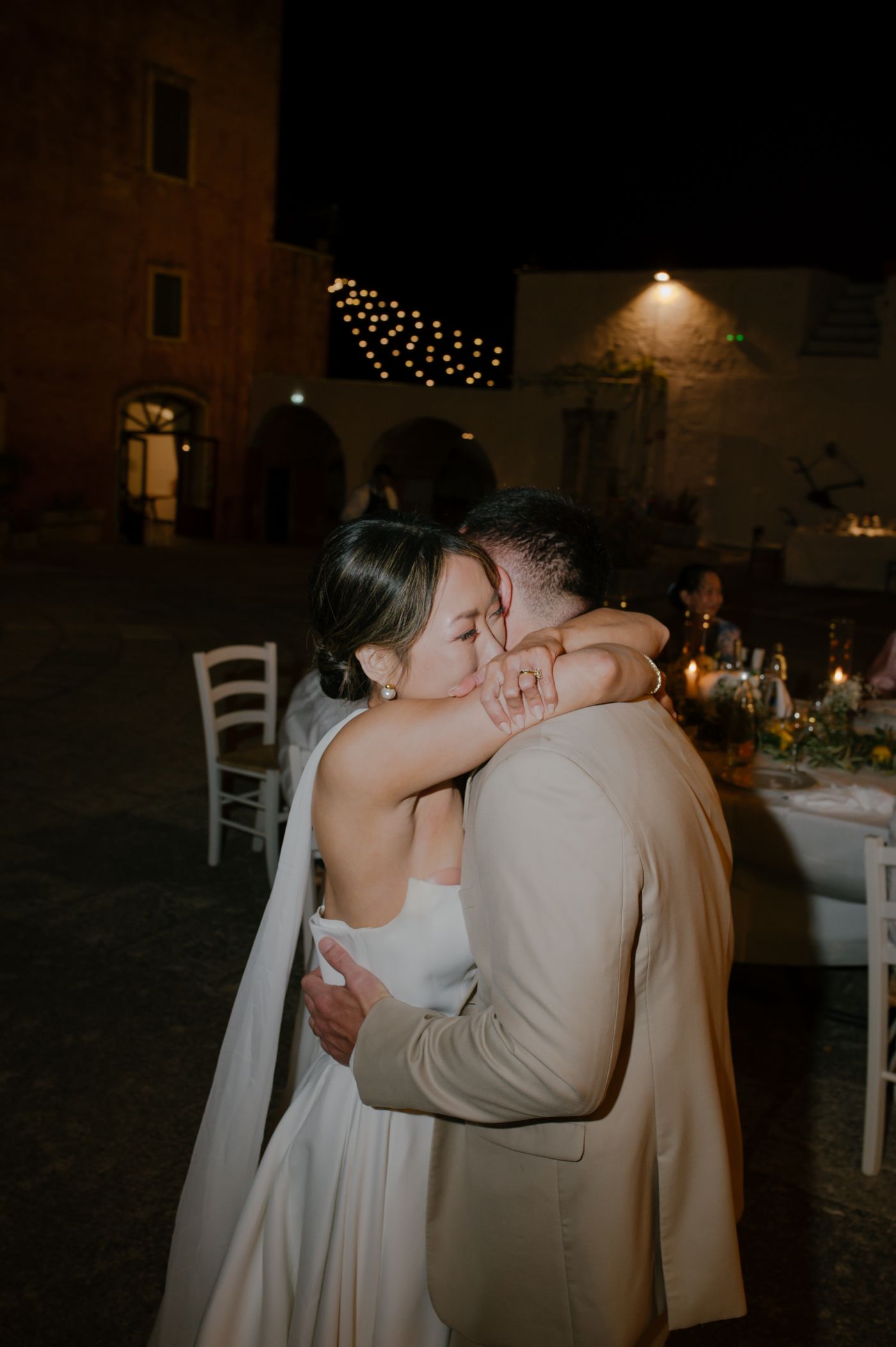 A romantic embrace between a couple at a dimly lit outdoor wedding reception with string lights overhead.
