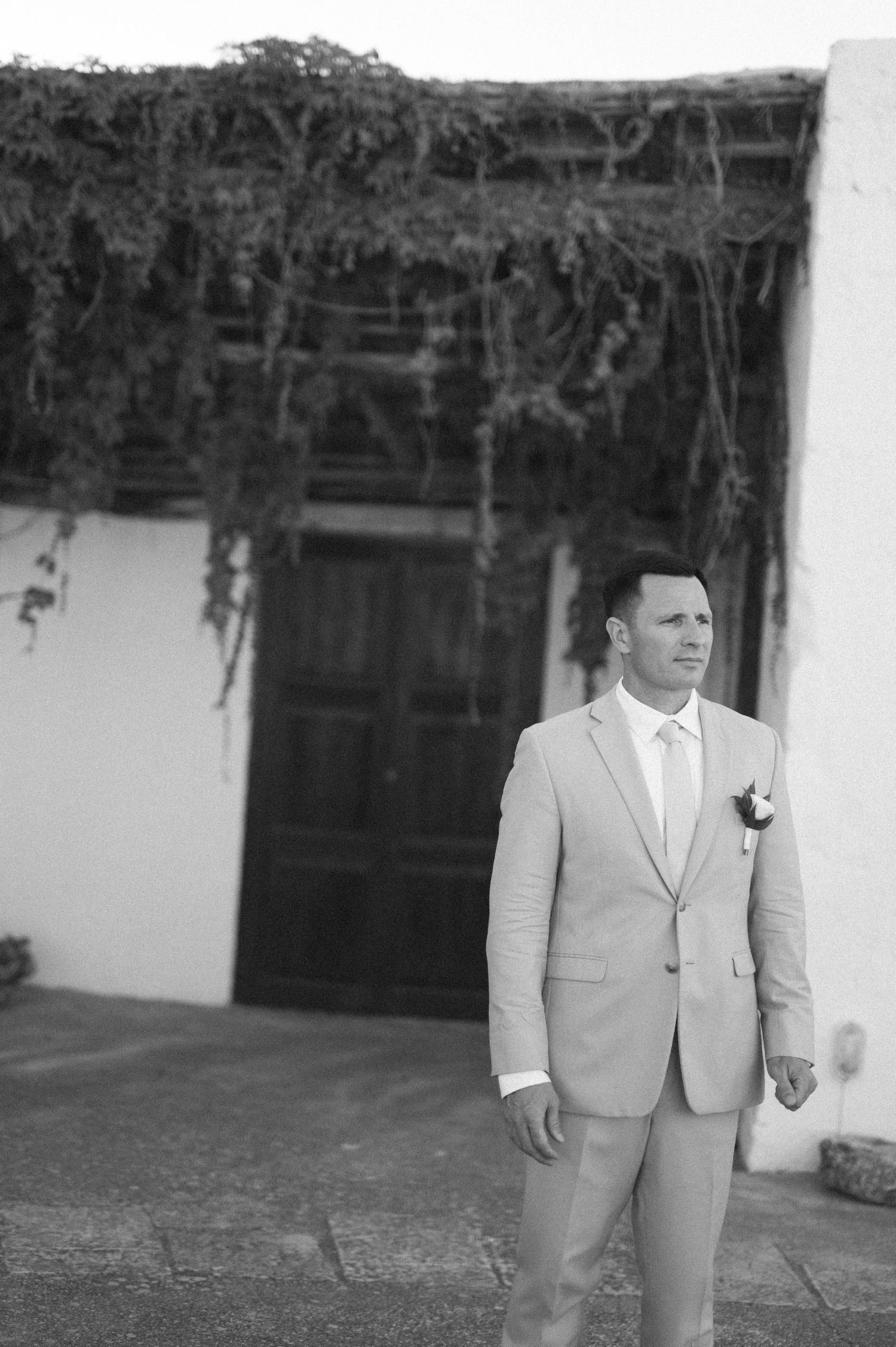 A groom in a light gray suit and white tie stands in front of a rustic doorway with hanging vines.