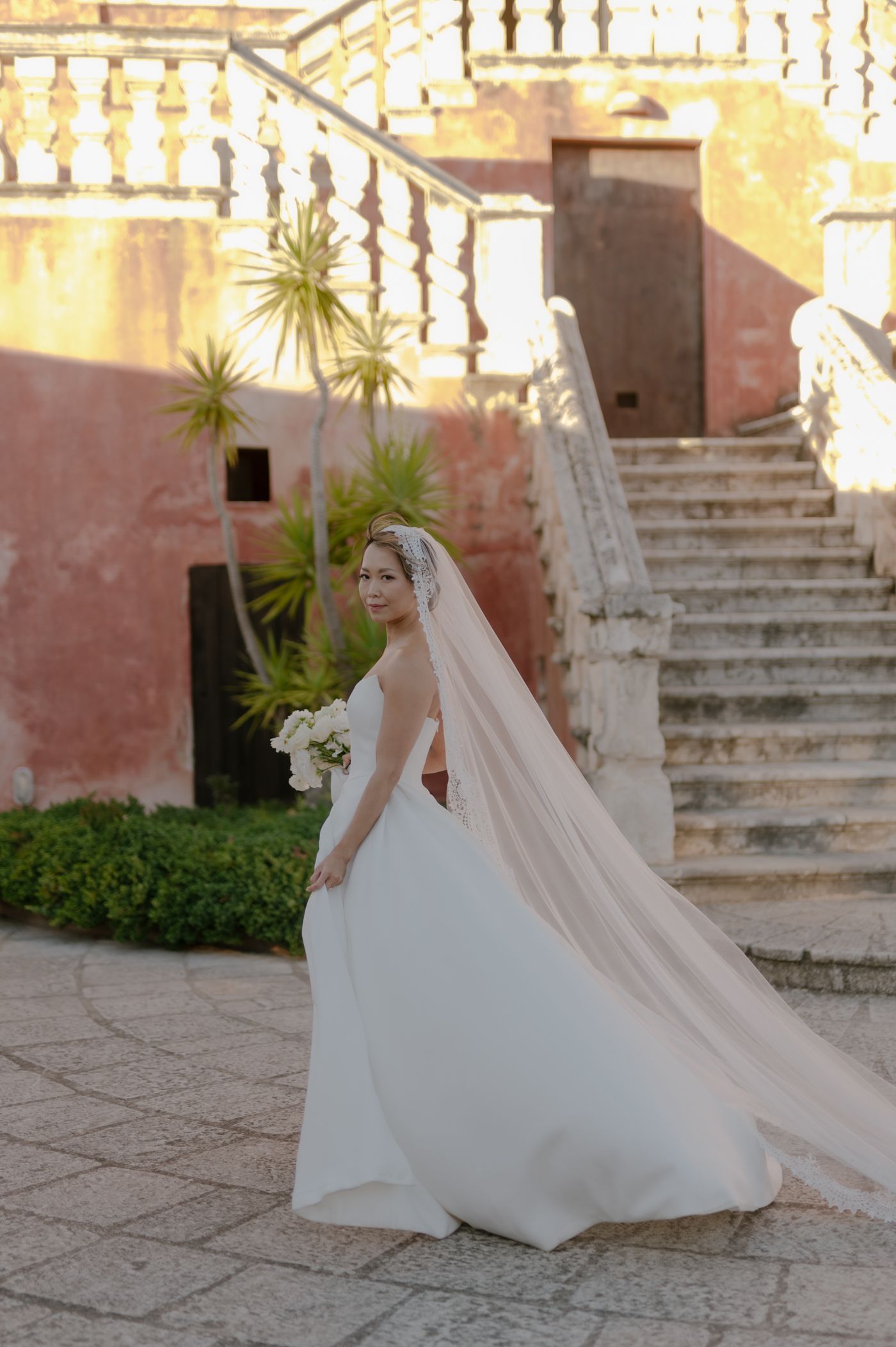 A flowing white wedding dress cascades down stone steps in a Mediterranean courtyard with pink walls and palm trees.