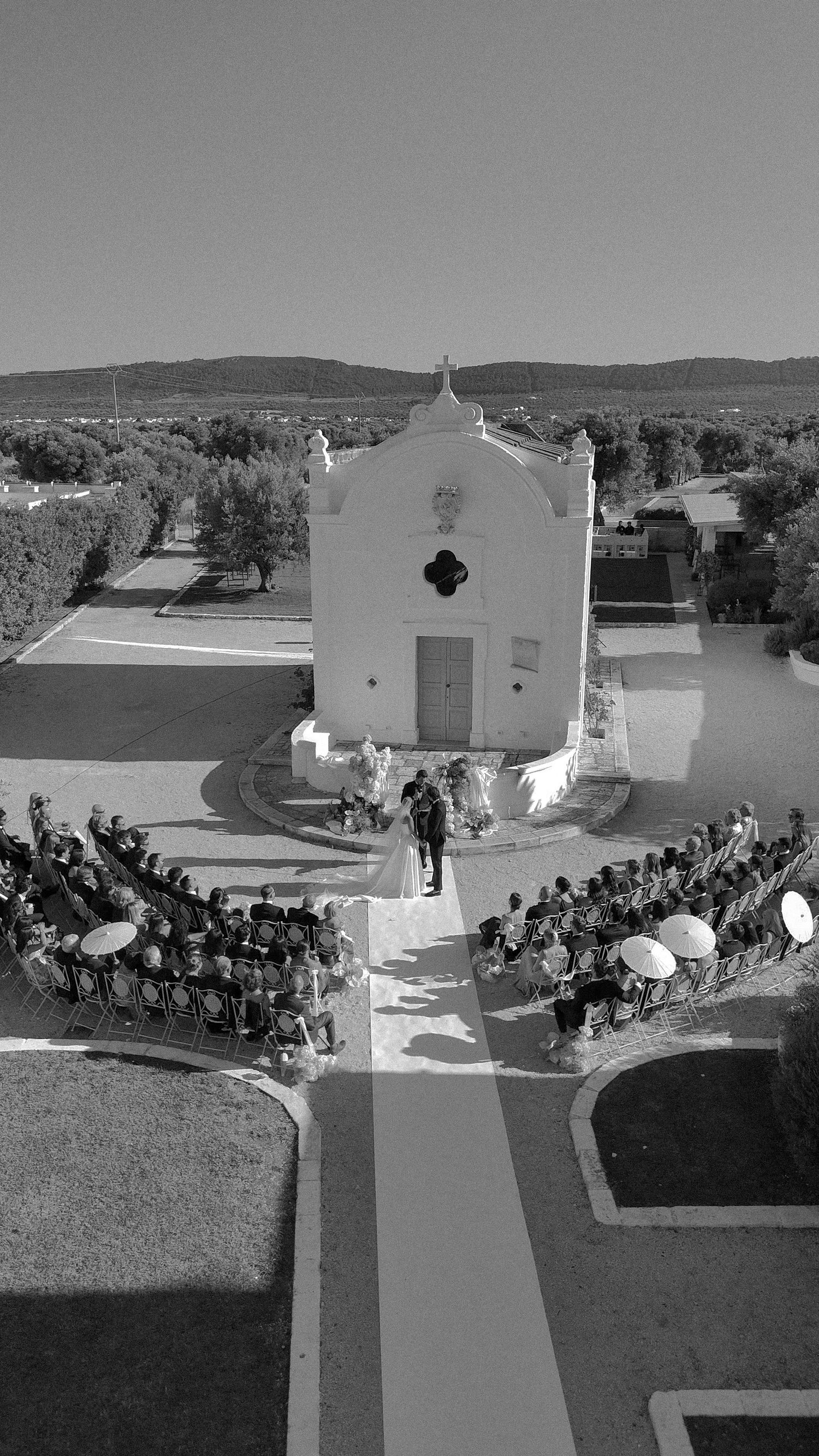 Aerial black and white photo of a wedding ceremony outside a white chapel surrounded by guests seated in rows.