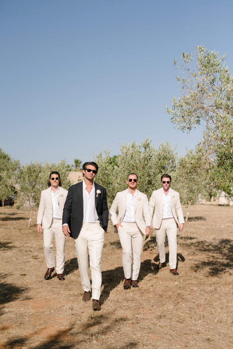Groomsmen in light colored suits walking through an olive grove.