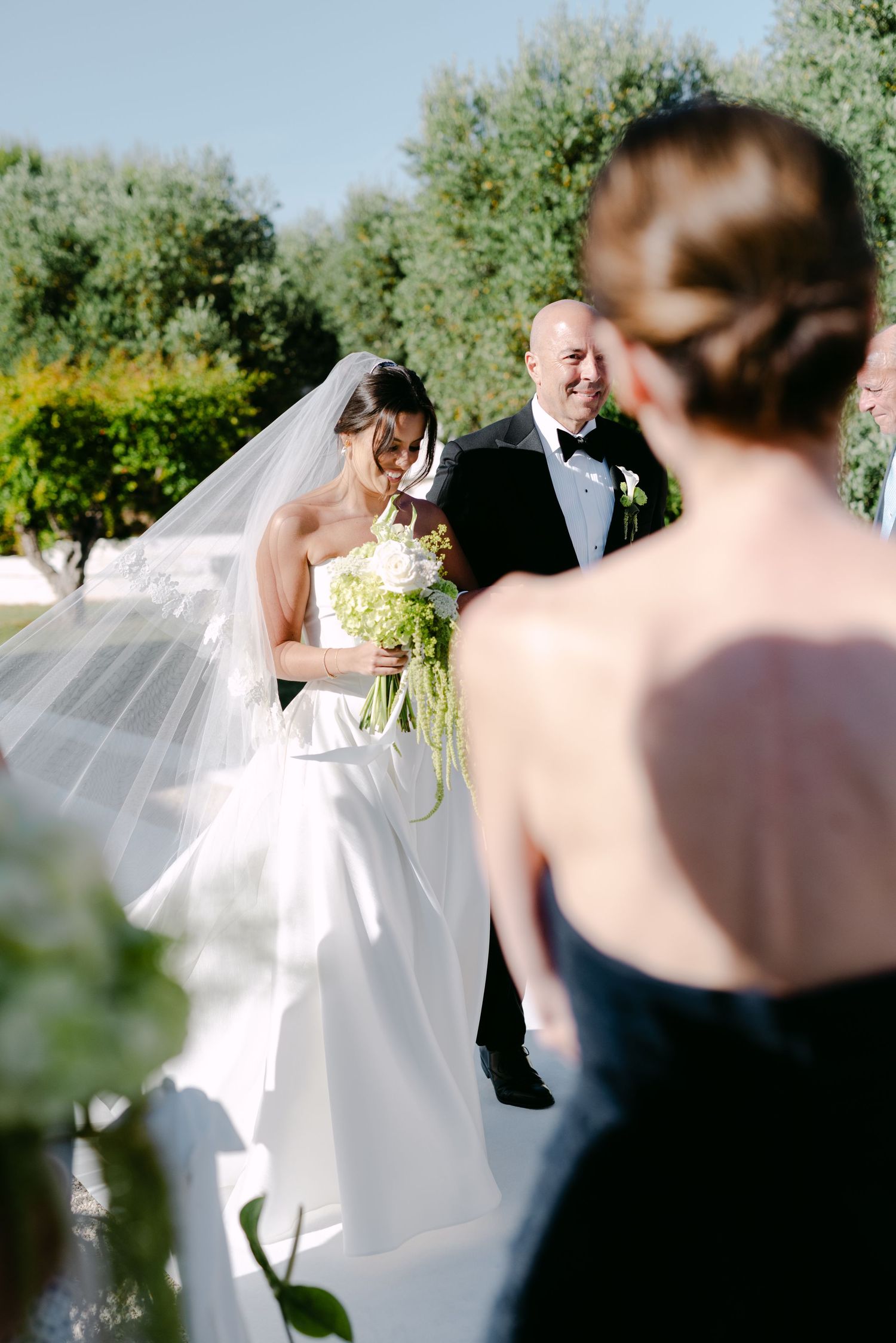 Bride in white gown and veil holds green bouquet beside groom in tuxedo at outdoor wedding ceremony.