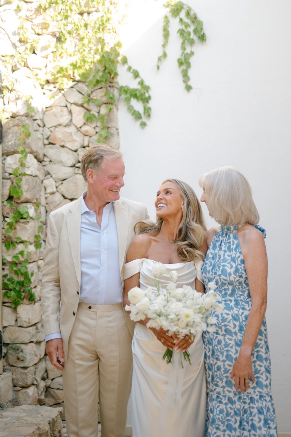 Wedding party sharing joyful moments against a white stone wall with climbing vines.
