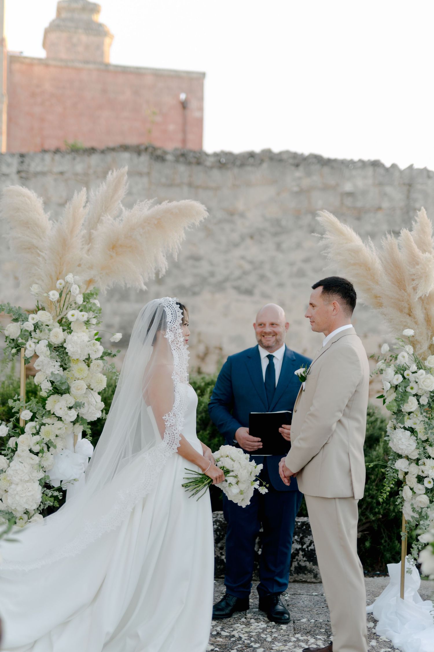 A wedding ceremony takes place outdoors with white floral arrangements and a veil flowing in the breeze.