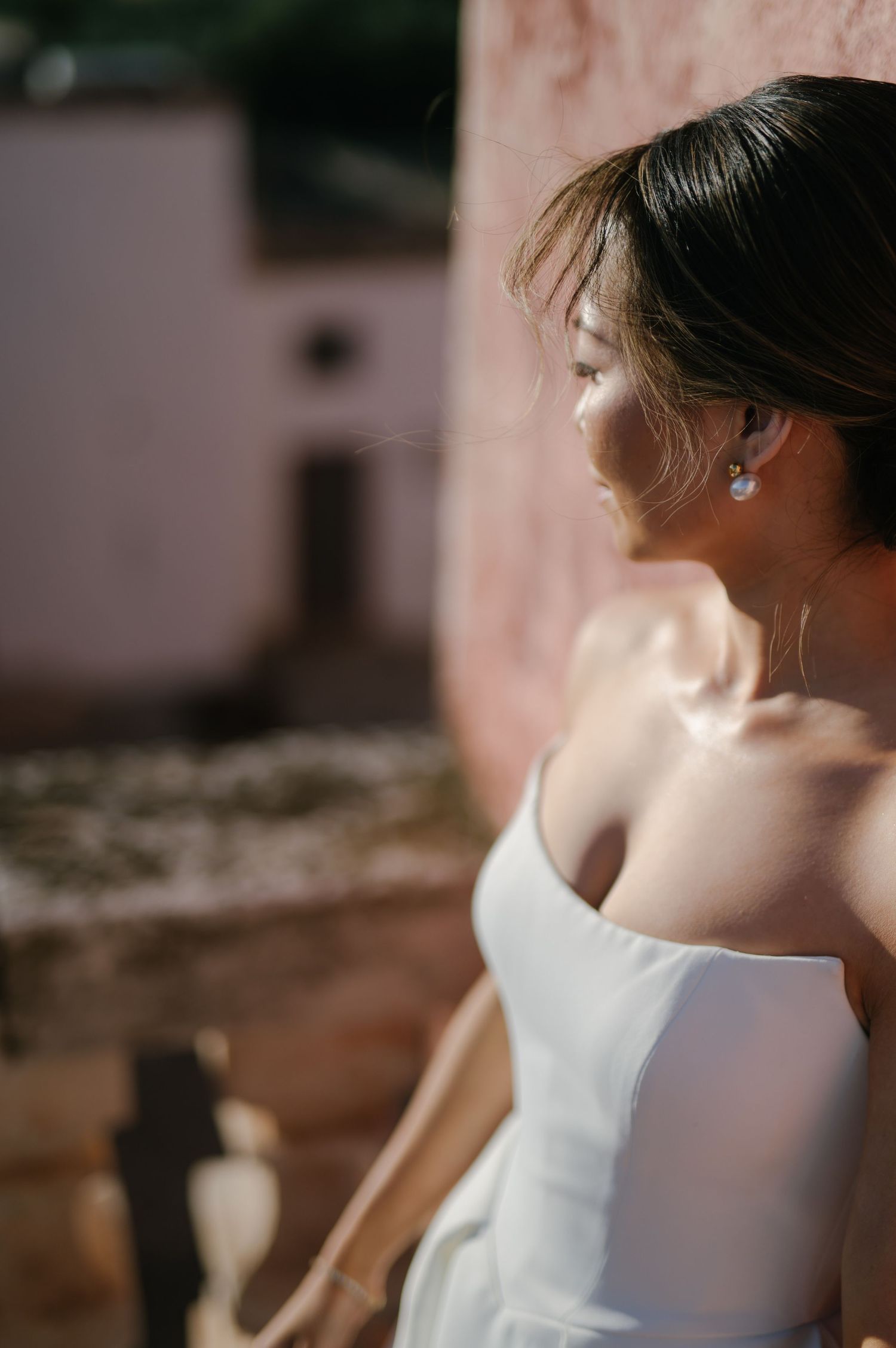Side profile of someone in a white off-shoulder dress with sunlit glow against a pink wall background.