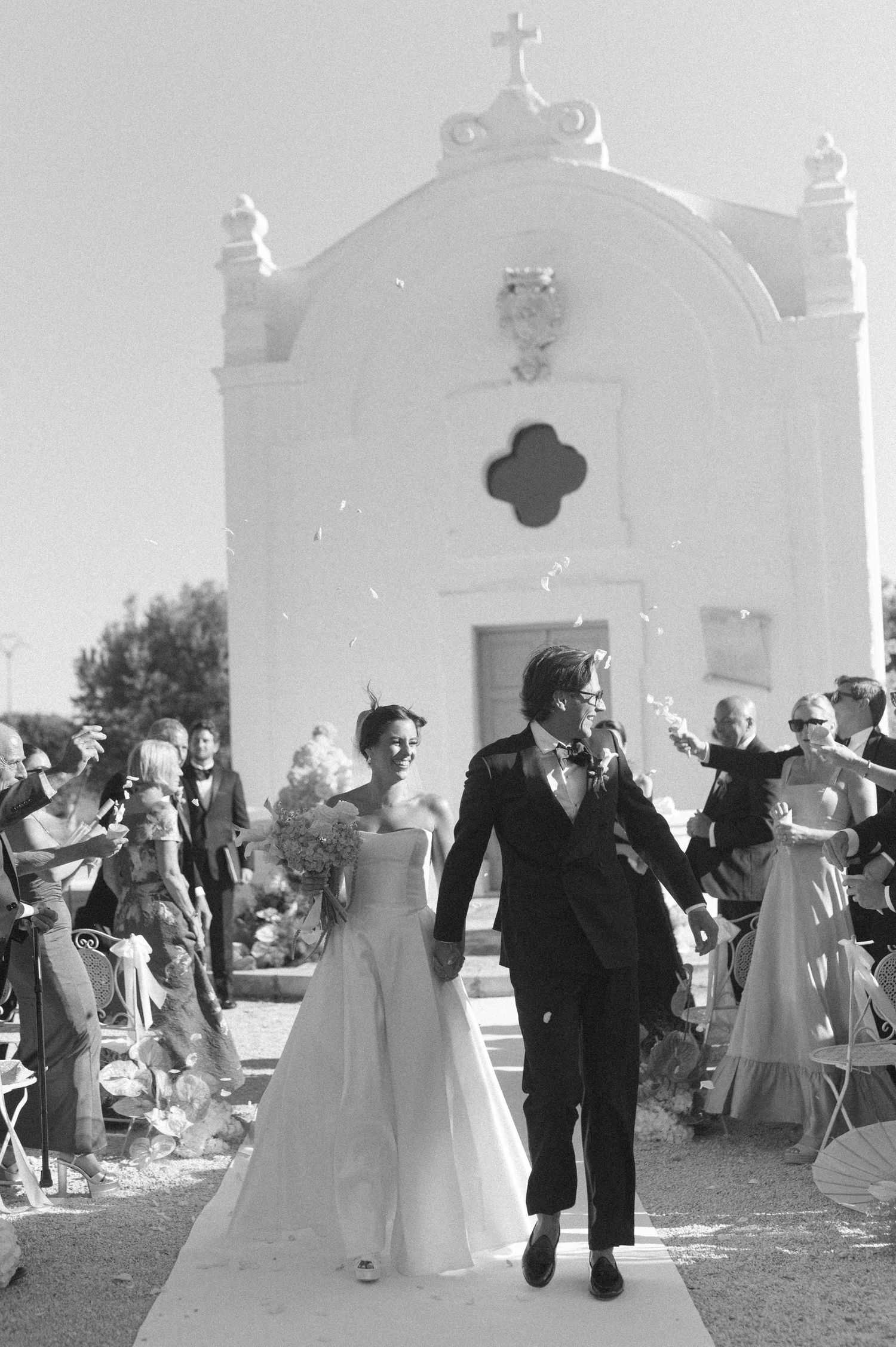 Bride and groom exit white chapel after wedding ceremony as guests celebrate with confetti in black and white photo.