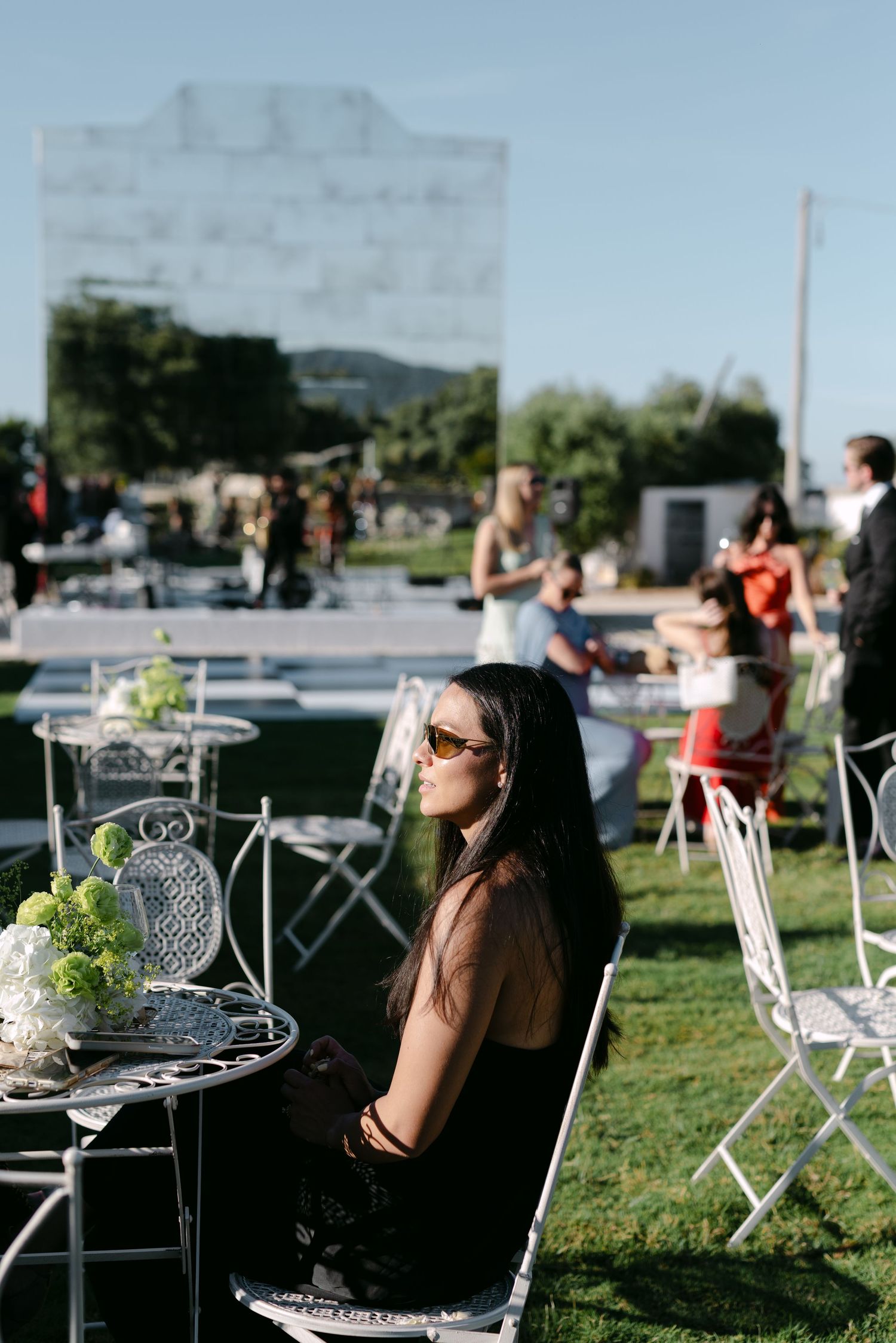 Woman in sunglasses sitting at ornate white garden table during outdoor wedding reception on sunny day.