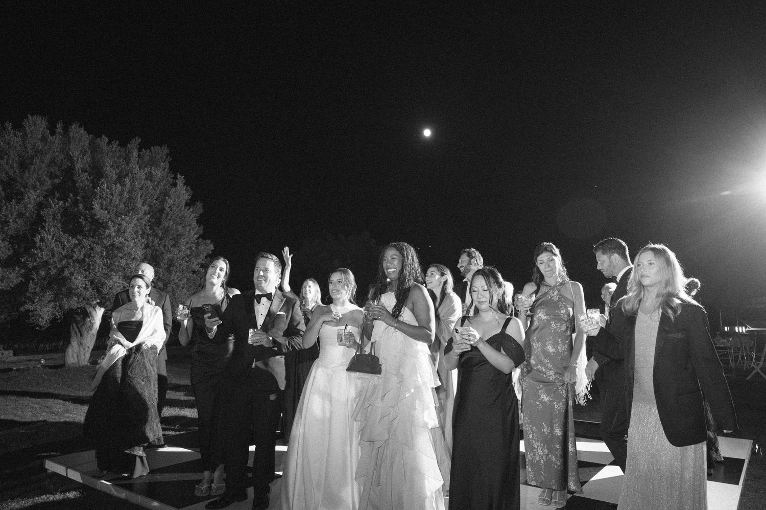 Black and white wedding party photo outdoors at night with bride and guests looking up at the moon.
