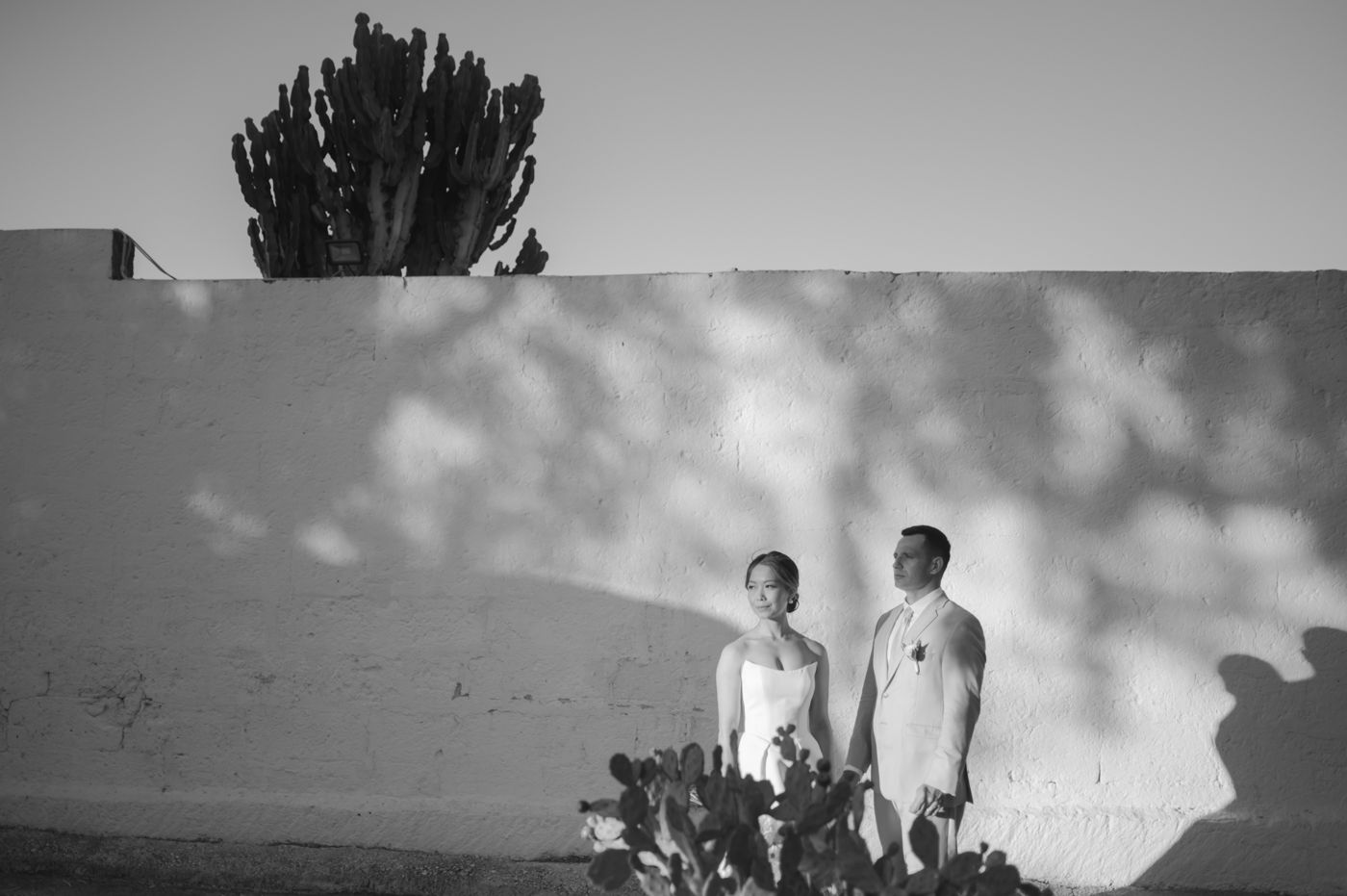 Black and white photograph of two figures standing against a white wall with dramatic shadows and a cactus silhouette.