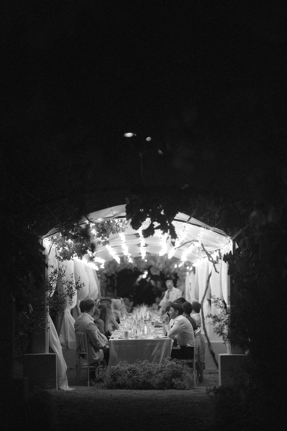 Silhouettes of wedding guests mingle under a romantically lit outdoor tent at night.