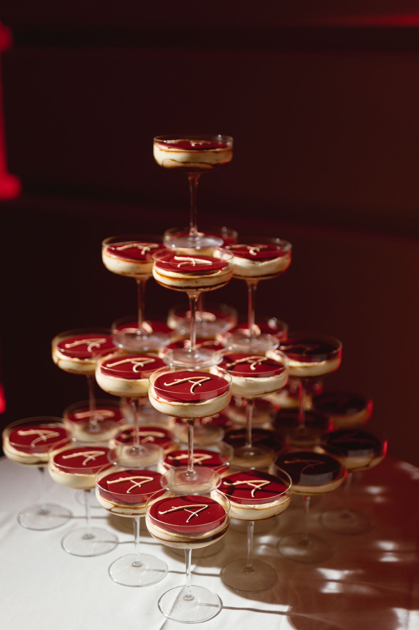 Red-tinted cocktails in coupe glasses arranged in elegant pyramid formation on reflective surface in dark ambient lighting.