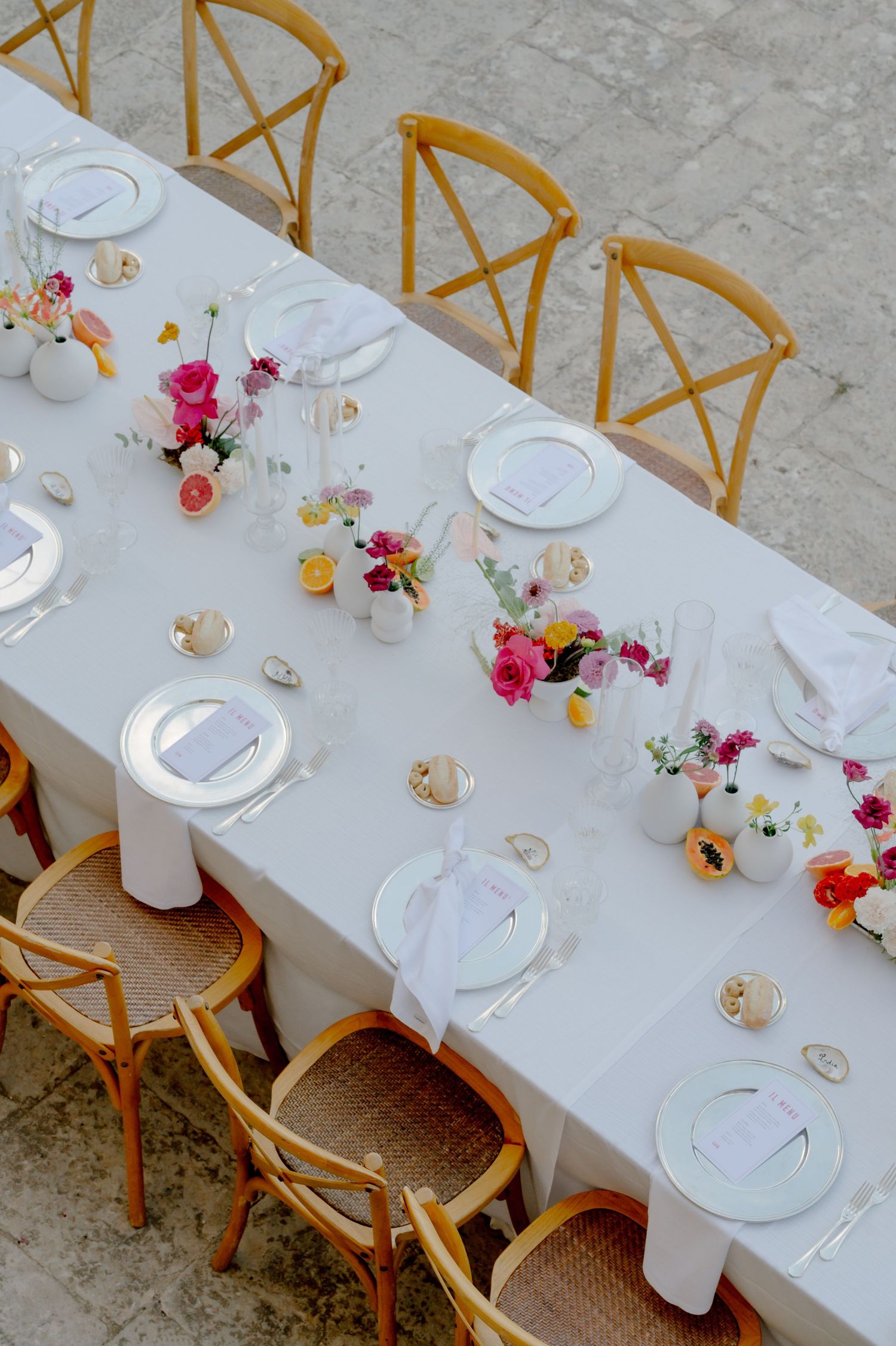 Long white dining table set with elegant place settings and pink floral centerpieces alongside wooden cross-back chairs.