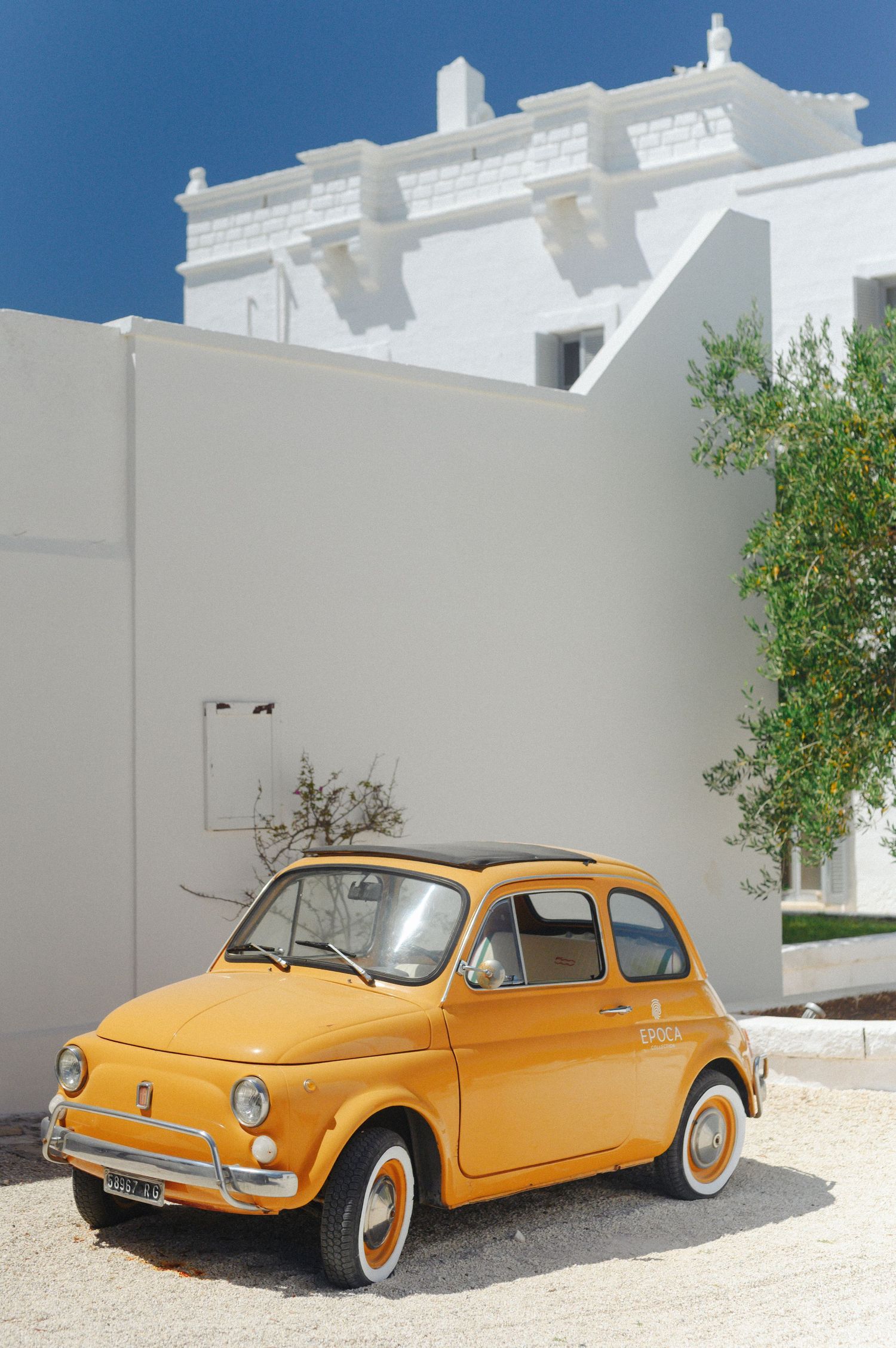 Vintage yellow Fiat 500 parked beside a white Mediterranean building under a clear blue sky.