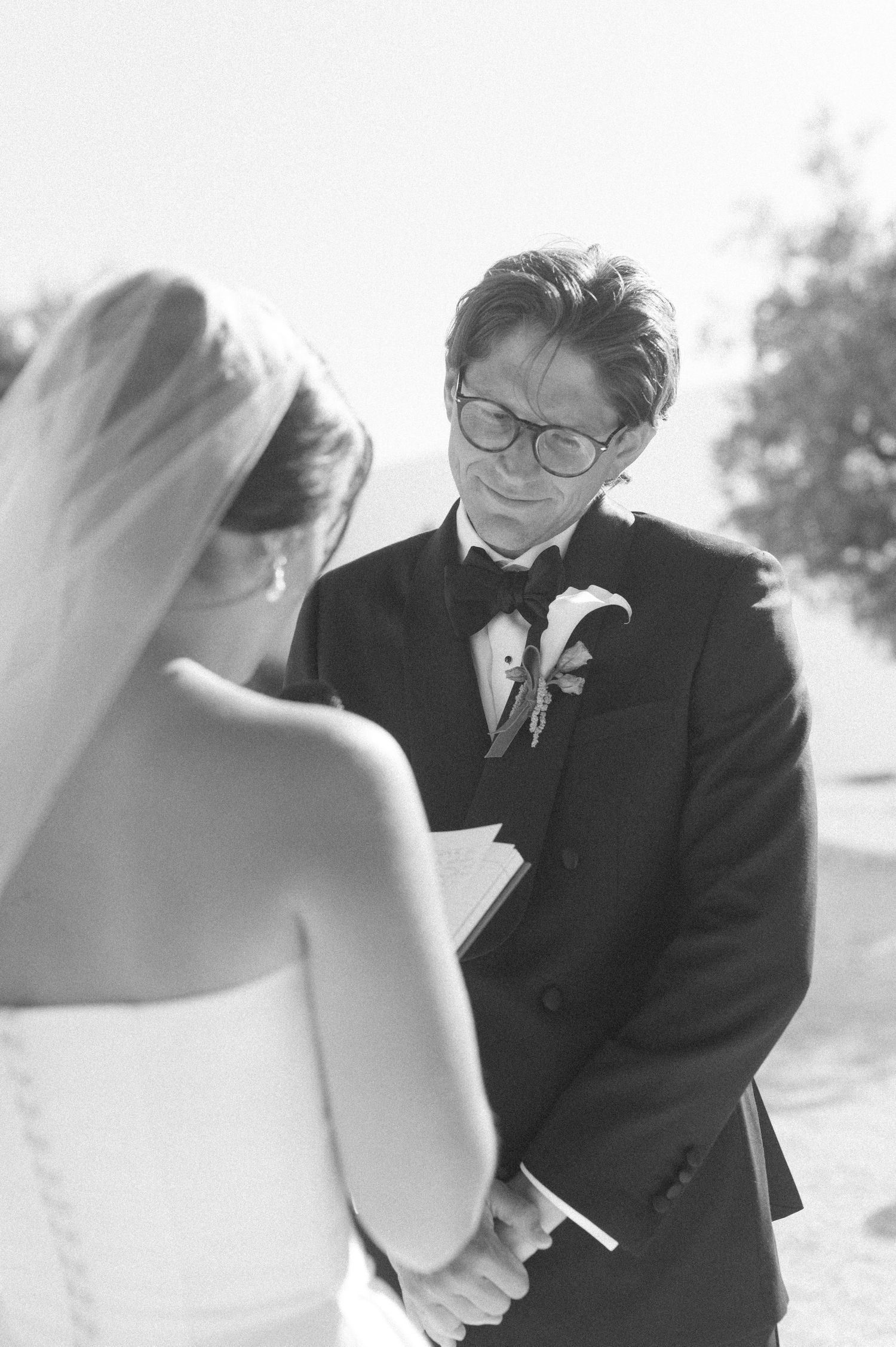 Black and white photo of a groom in a tuxedo reading vows to his bride during an outdoor wedding ceremony.