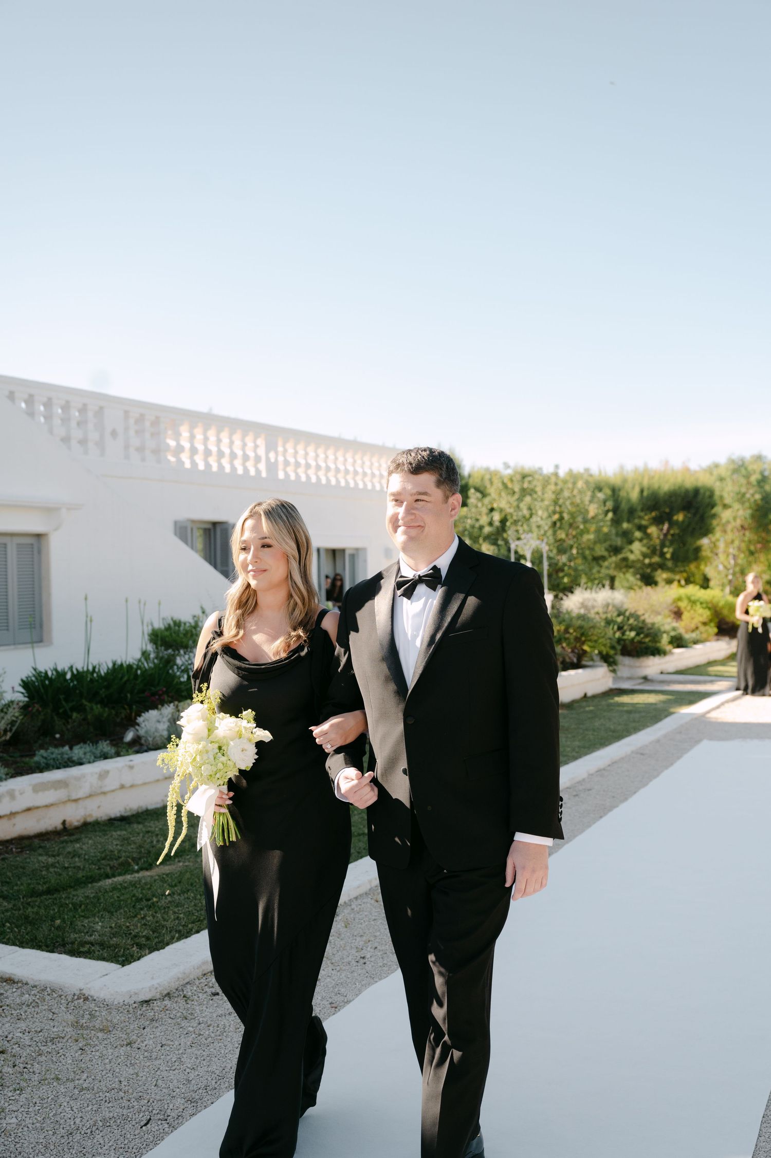 A couple walks arm in arm in black formal attire, woman holding white bouquet, at an elegant outdoor wedding venue.
