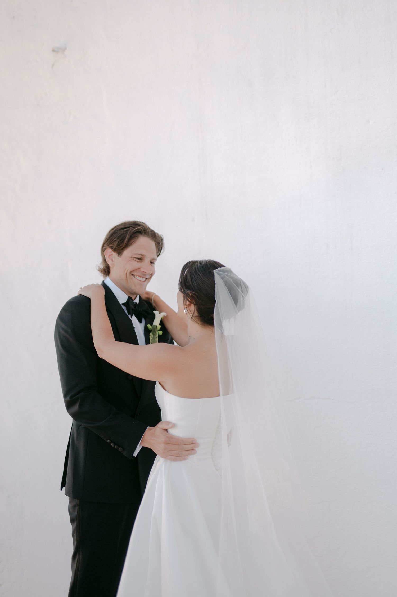 Smiling groom in black tuxedo embraces bride in white wedding dress and veil against a white wall.