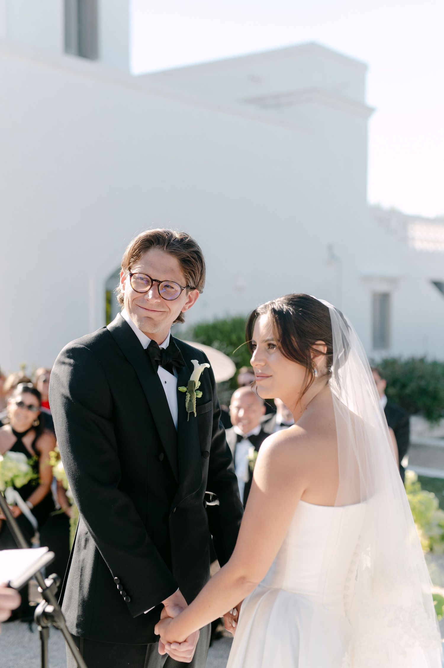 A bride and groom hold hands during an outdoor wedding ceremony, dressed in a white gown and black tuxedo.