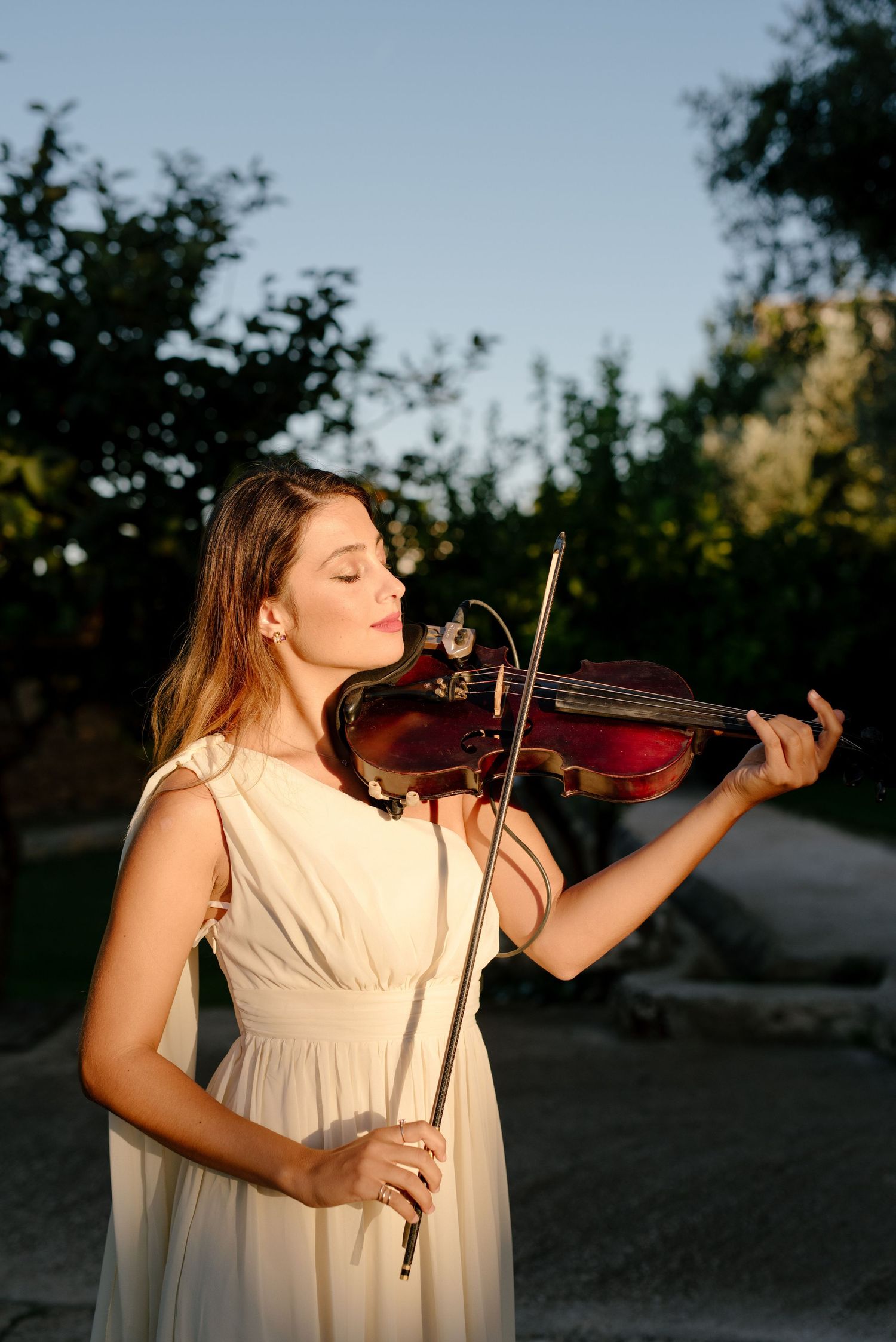 A violinist in a white dress performs outdoors at sunset, creating a serene and artistic atmosphere.
