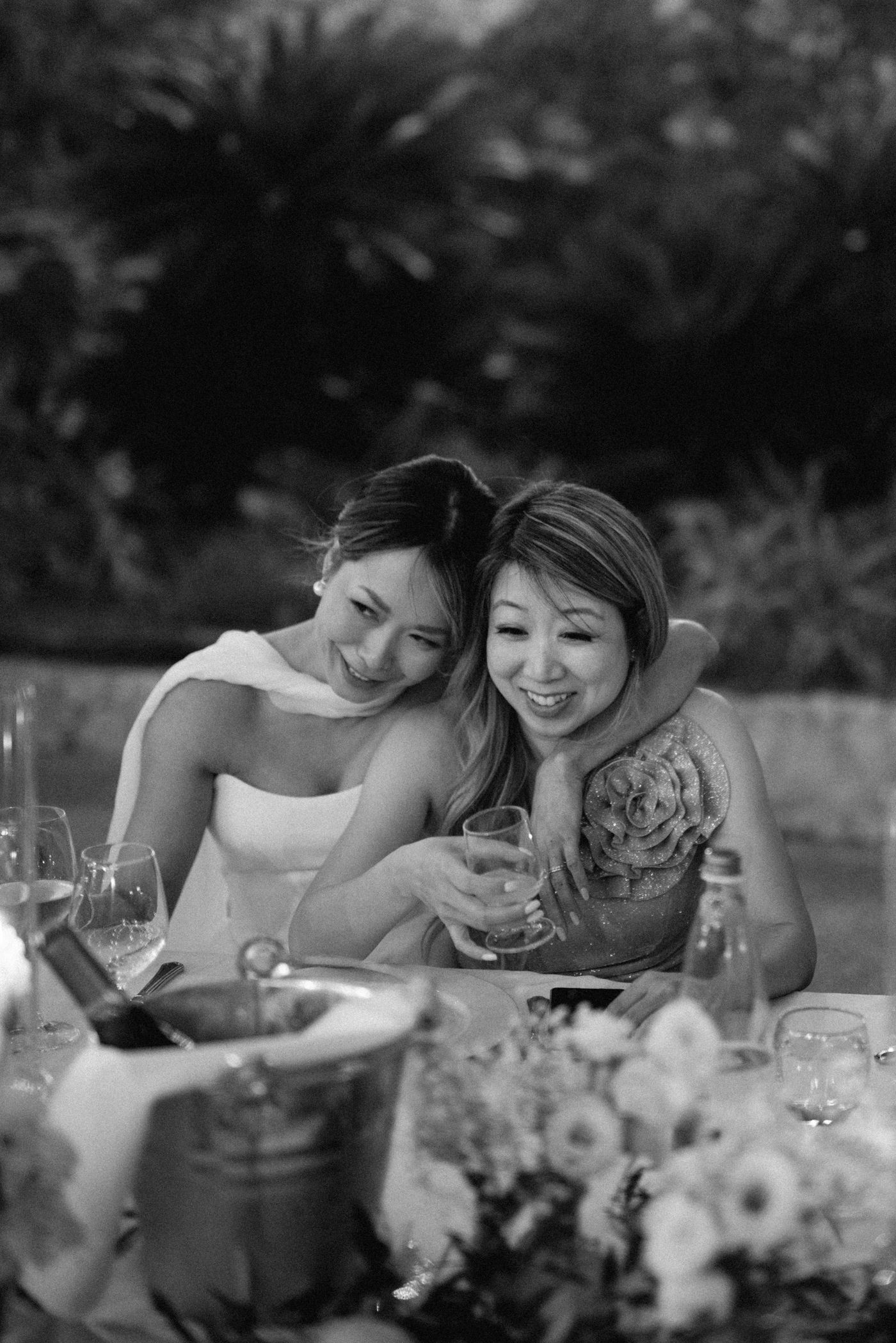 Black and white photo showing two people laughing together at an elegant dinner table with floral centerpieces.