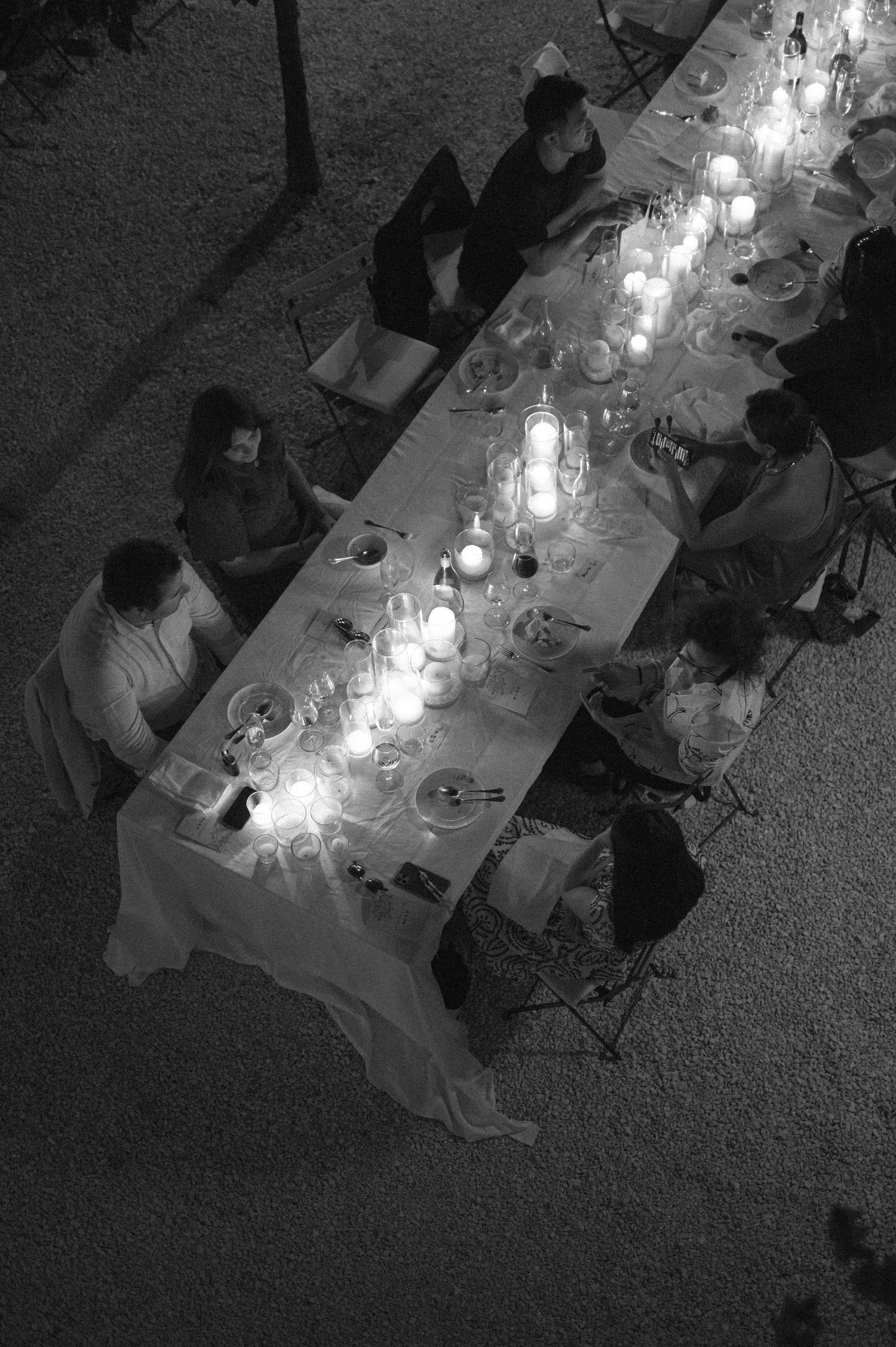 Overhead view of a candlelit dinner table with guests seated at a long white-clothed table in a dark room.