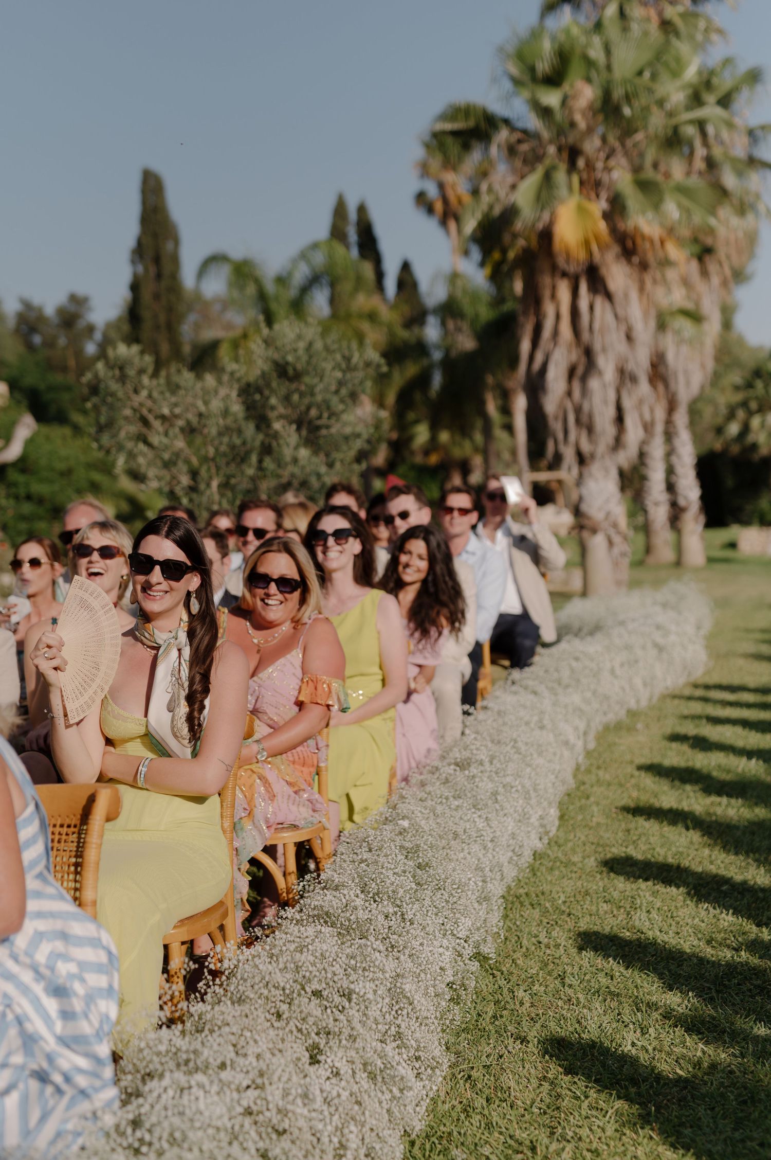Wedding guests wearing sunglasses sit in rows of white chairs along a palm-lined path during an outdoor ceremony.