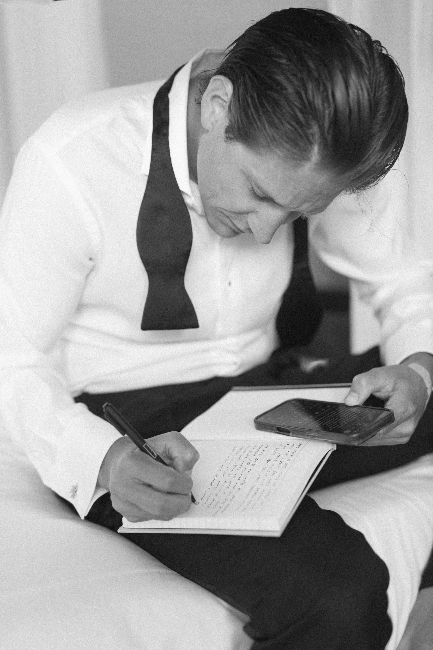 Man in white dress shirt and bow tie writing in notebook while holding smartphone, black and white photo.