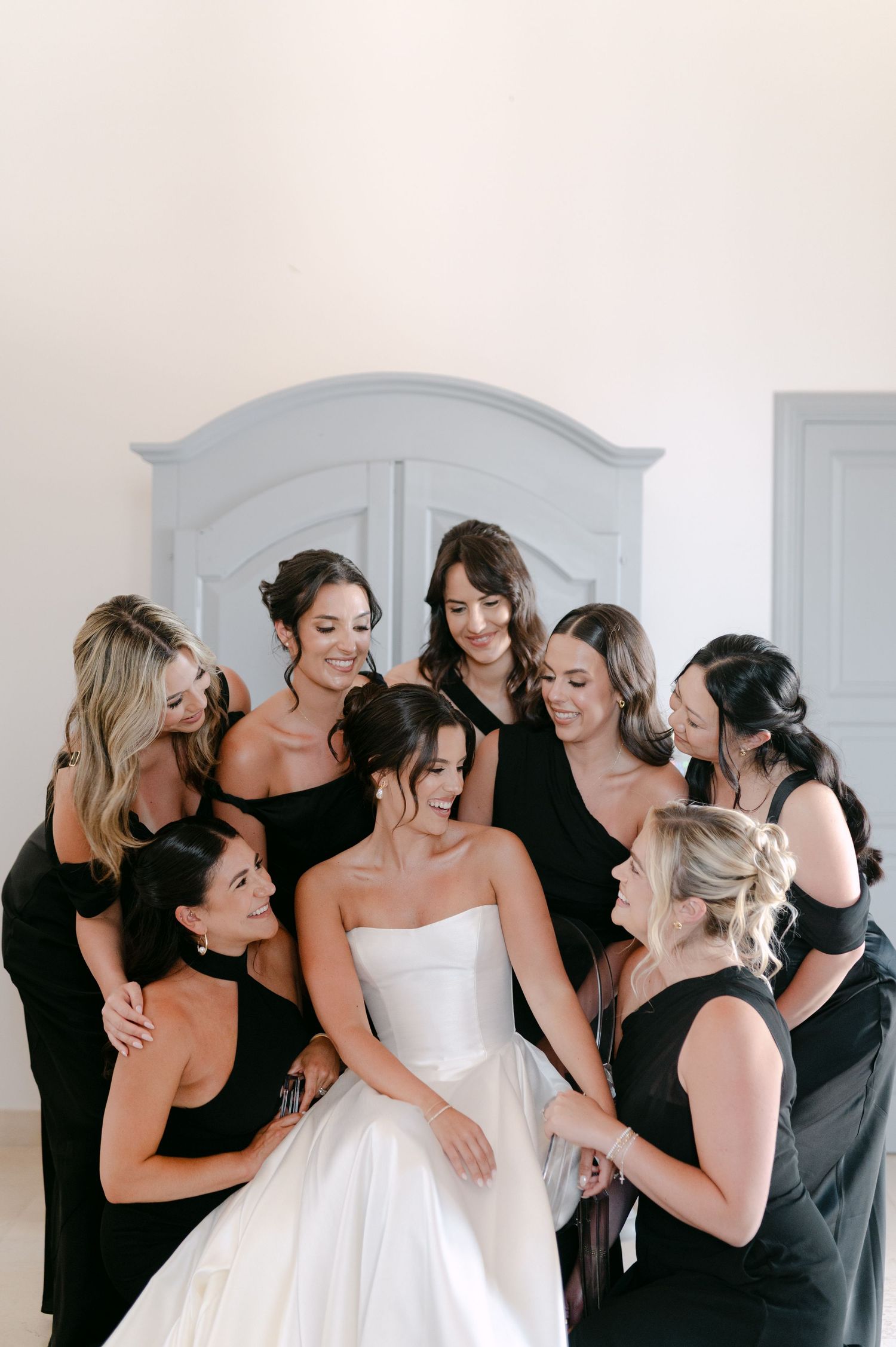 Bride in white gown surrounded by smiling bridesmaids in black dresses, sharing a joyful moment before the wedding.