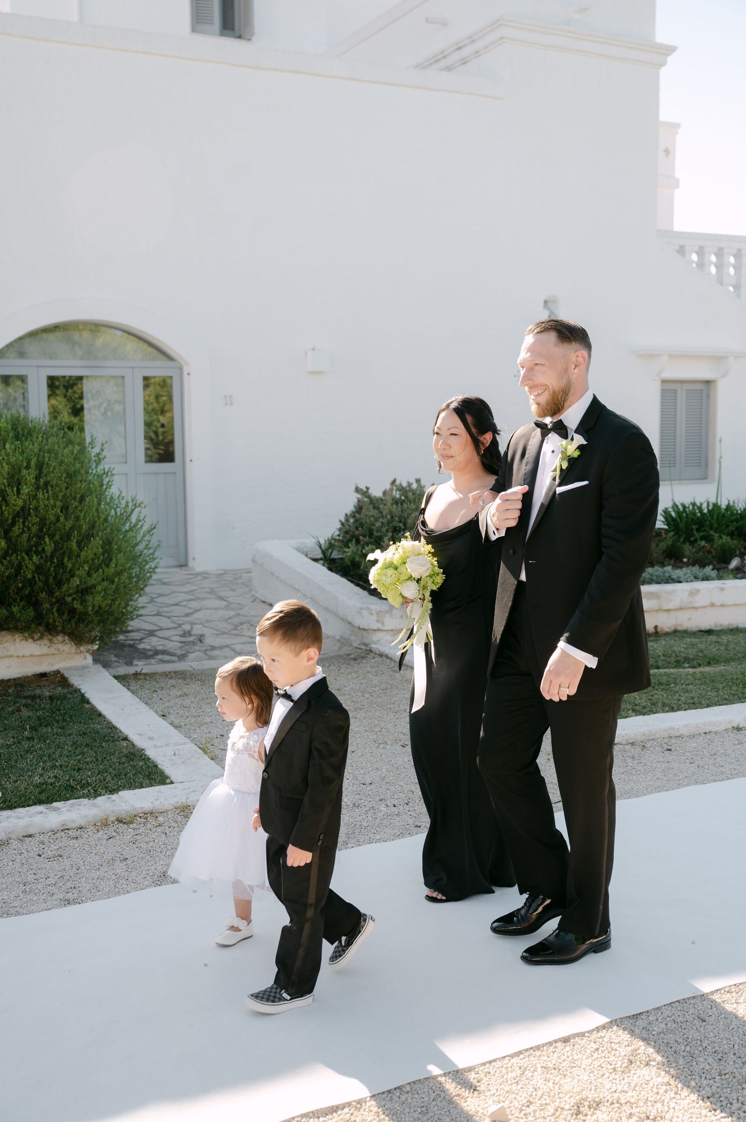 Bride and groom walk with two children in formal attire outside a white villa on their wedding day.