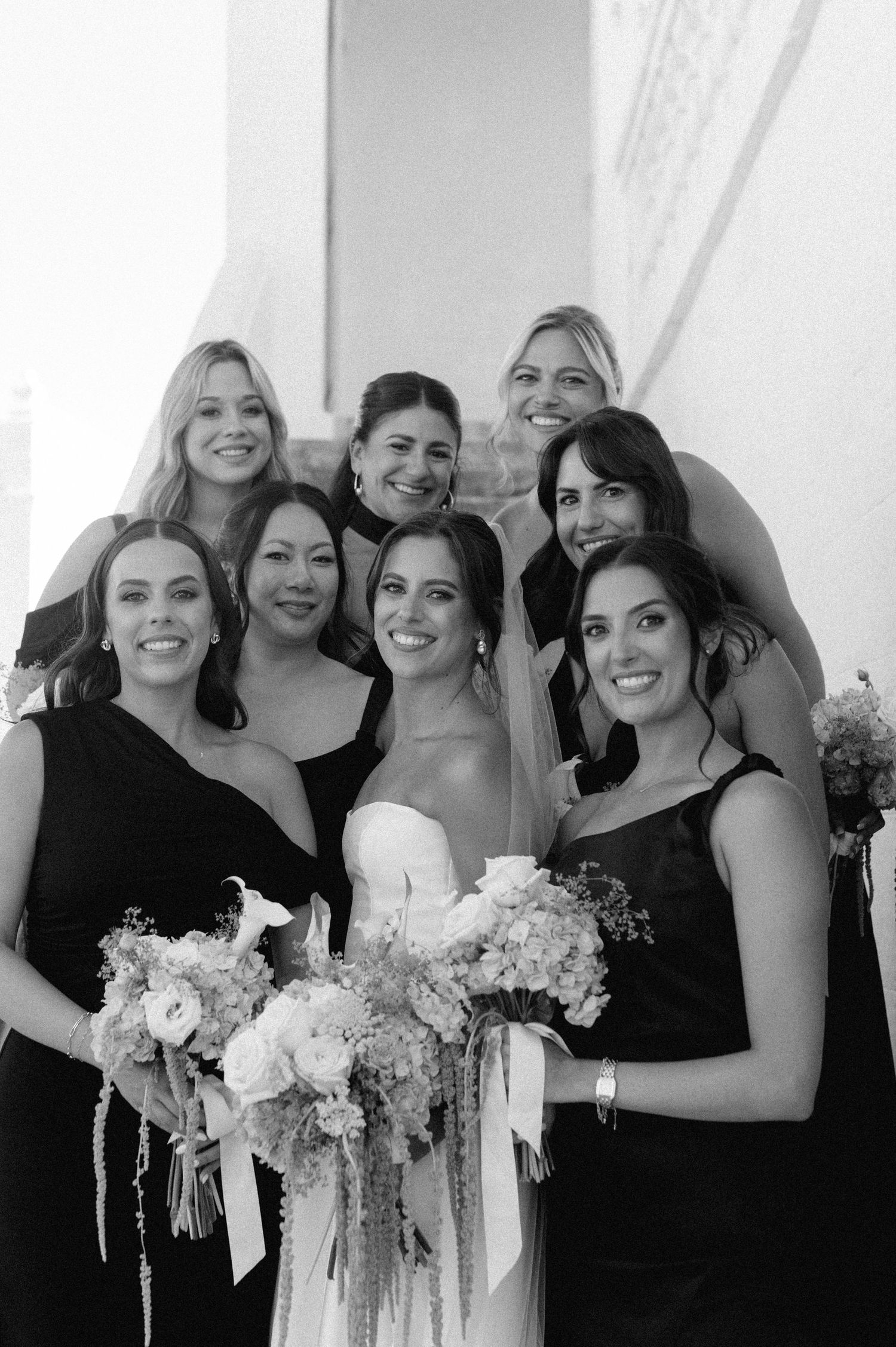 Black and white photo of a bride in a strapless gown with bridesmaids in black dresses holding floral bouquets.