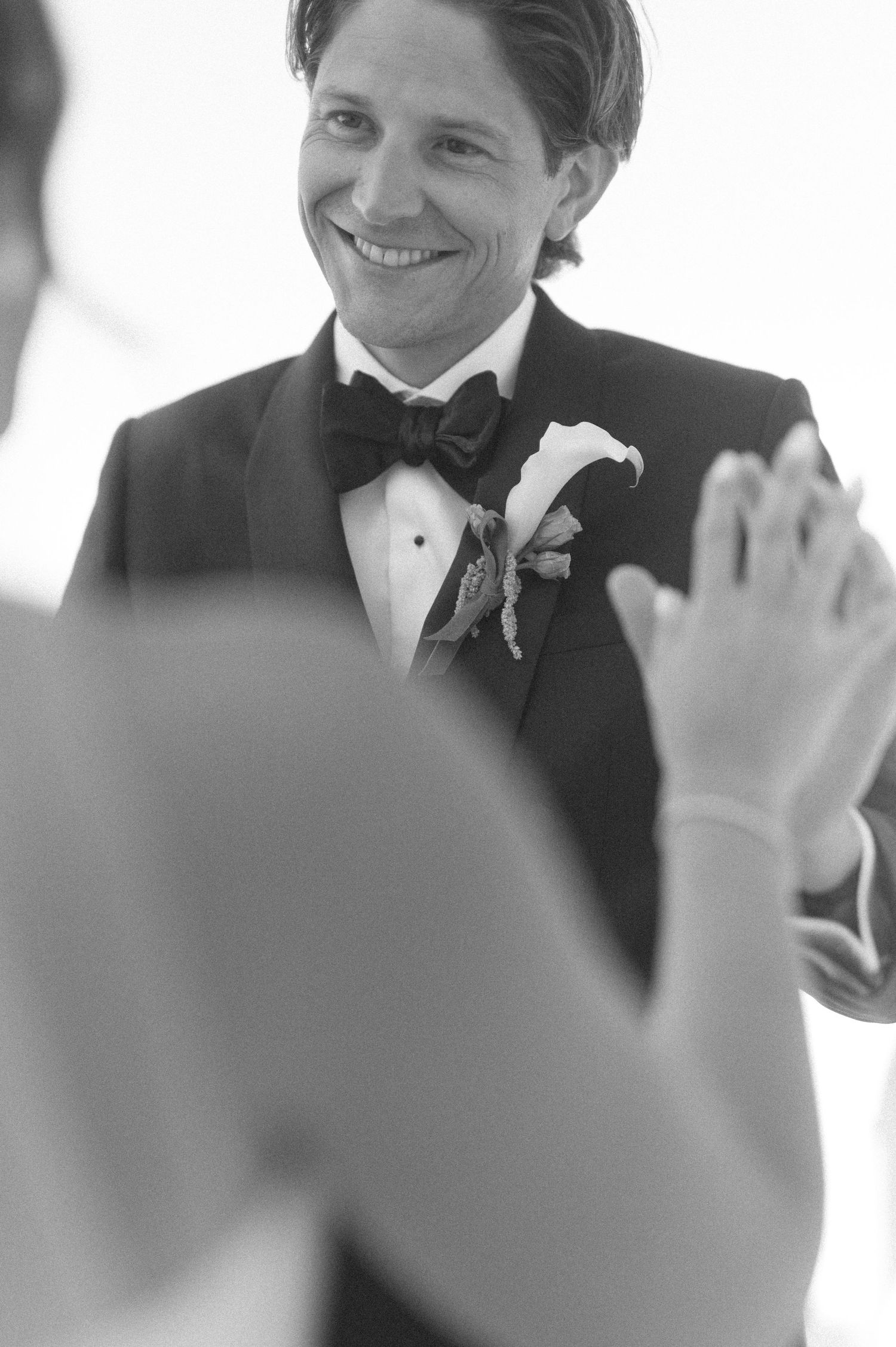 Smiling groom in black tuxedo with bow tie and floral boutonniere during wedding ceremony in black and white photo.