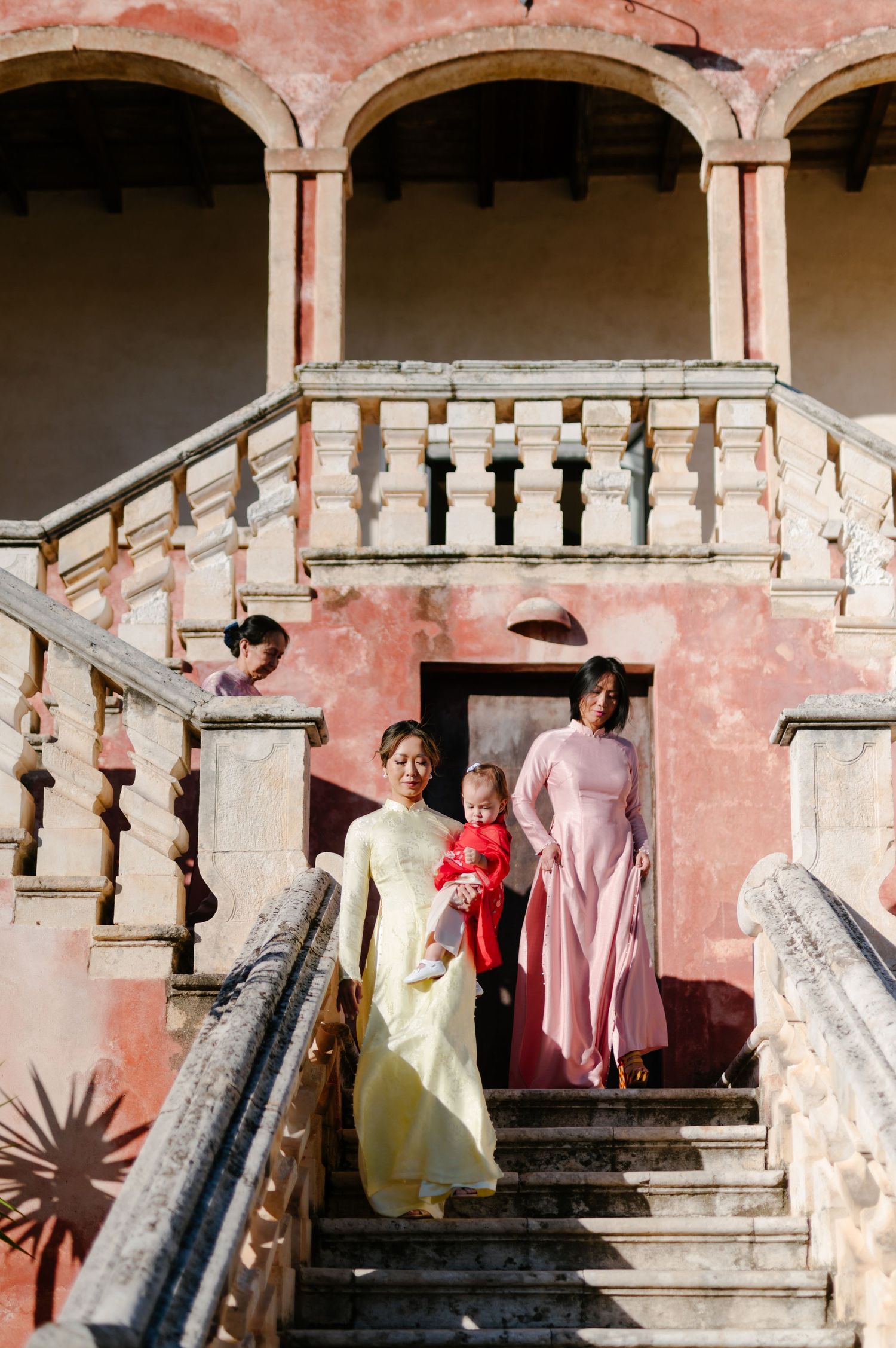 People in vintage-style dresses stand on a grand stone staircase with pink walls and arched columns in an elegant setting.