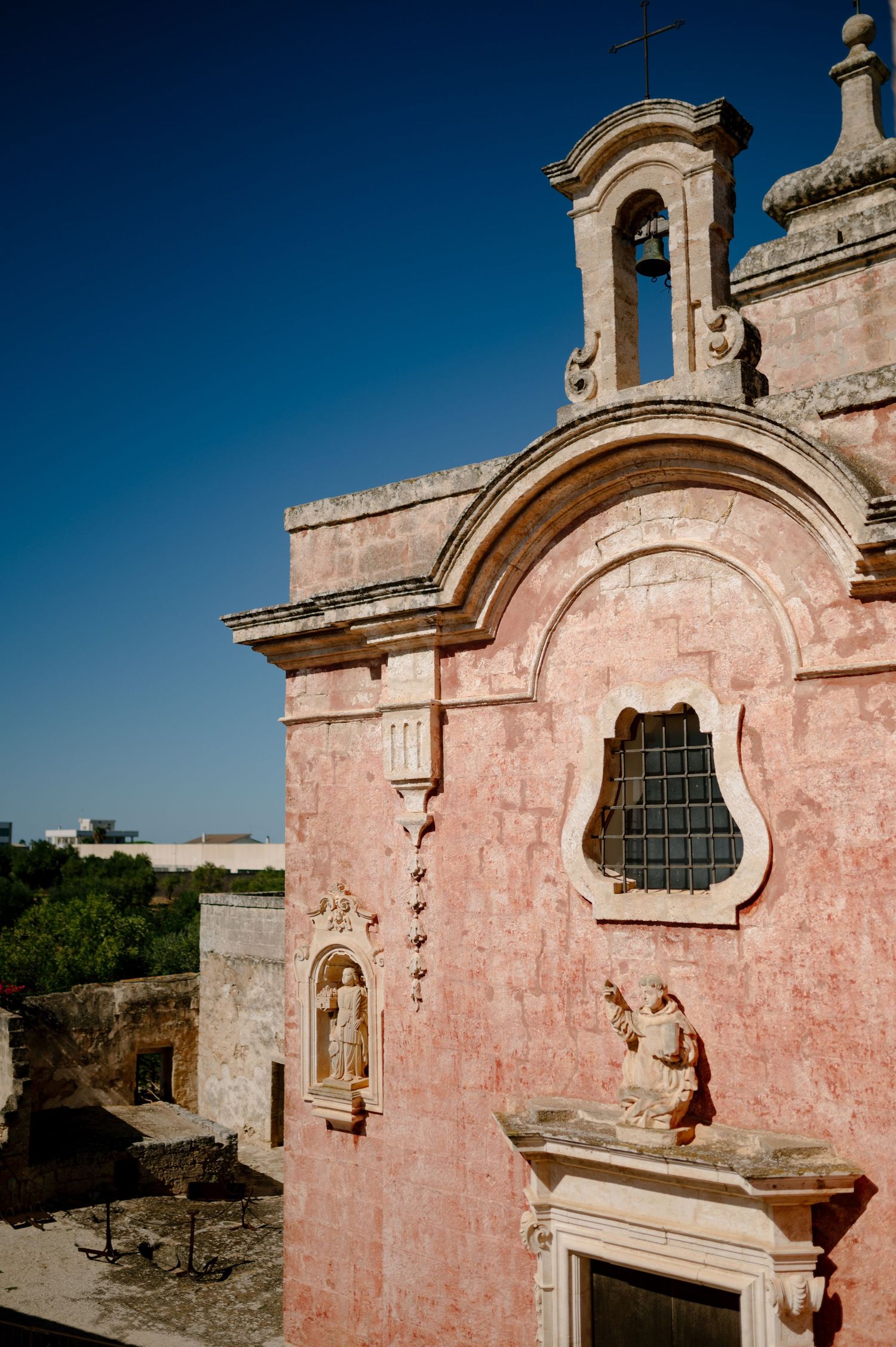 Pink stone church facade with ornate bell tower and arched window against bright blue sky in Mediterranean architectural style.