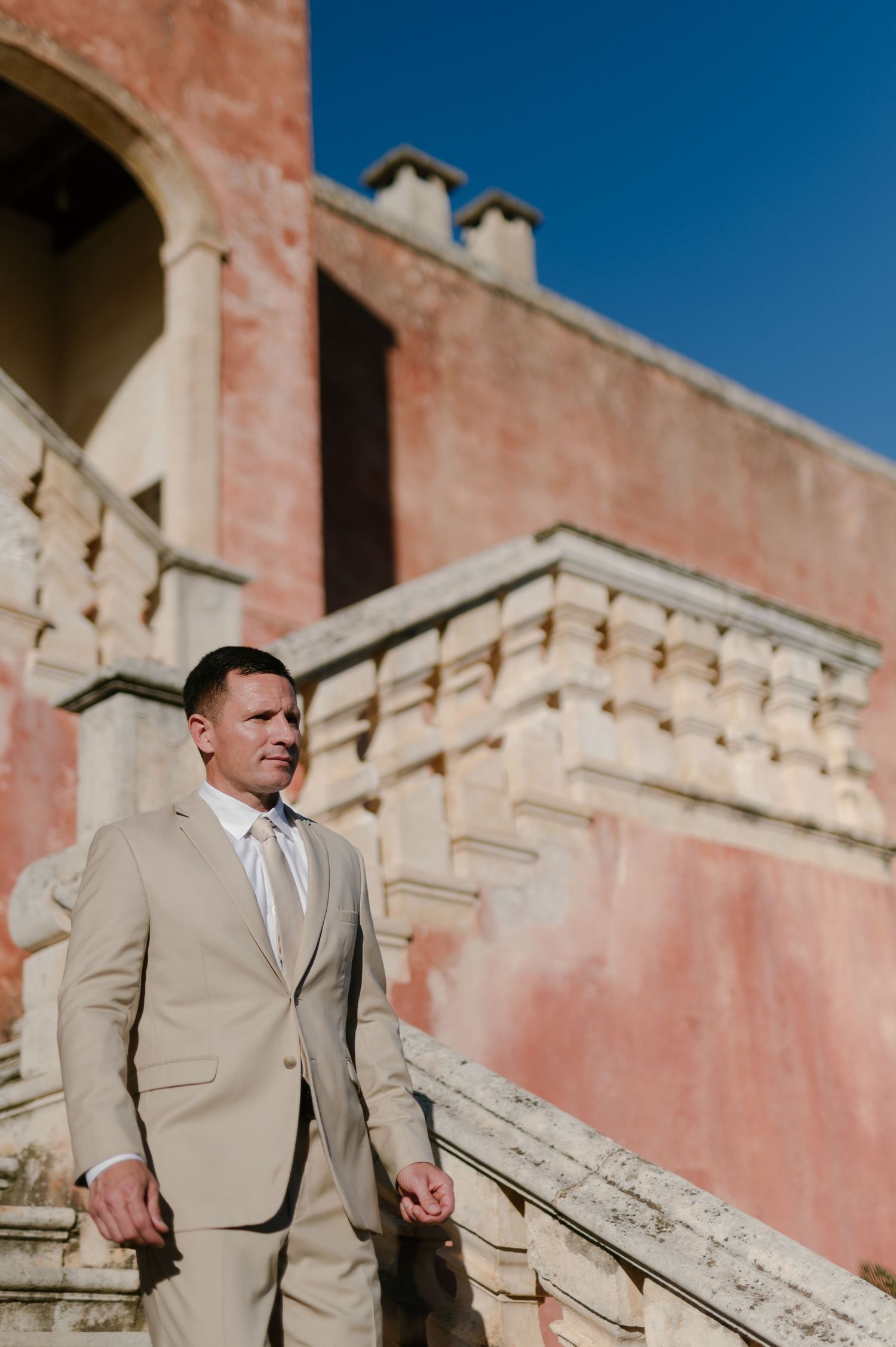 A groom in a beige suit poses against a pink stucco wall with architectural details on a sunny day.