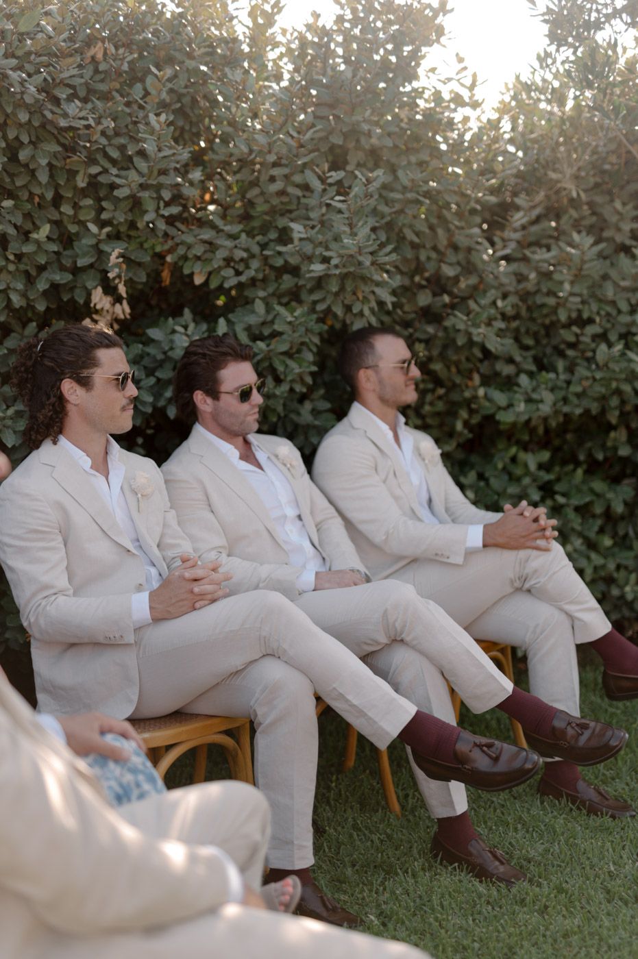 Groomsmen in white suits relaxing on chairs in garden setting.